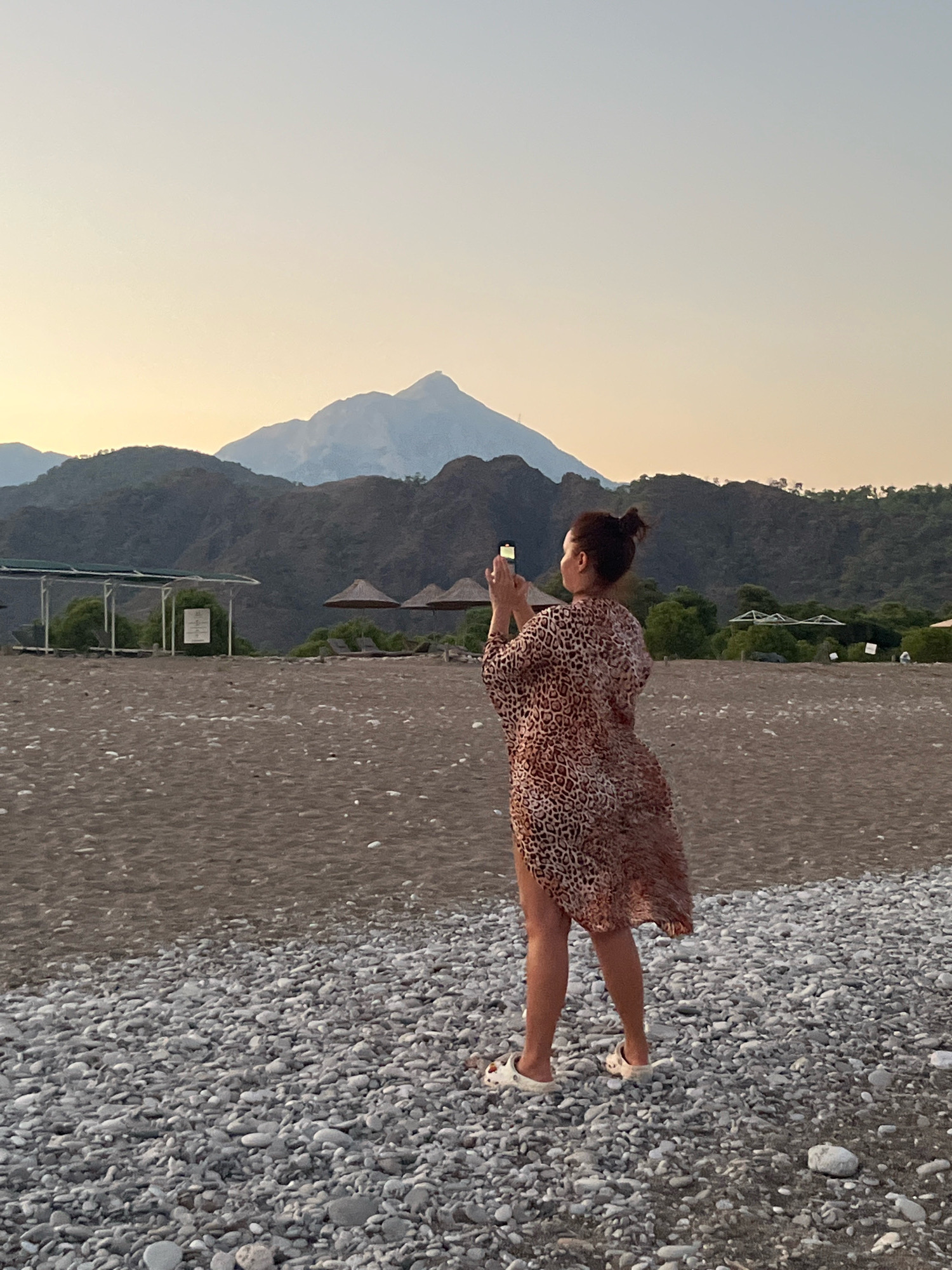 A full-shot image shows a woman standing on a pebble beach, facing away from the camera and towards a mountain range under a hazy, pale yellow sky. The woman has her hair pulled up in a bun and is wearing a leopard-print cover-up and white slip-on shoes. She is holding a smartphone in her hands, seemingly taking a photo of the landscape. In the background, there are beach umbrellas, lounge chairs, and scattered trees. The beach itself is a mix of sand and smooth, grey pebbles. The overall scene suggests a tranquil evening in a coastal area, possibly in or near Kemer, Turkey, given the mountainous terrain and resort-like amenities.