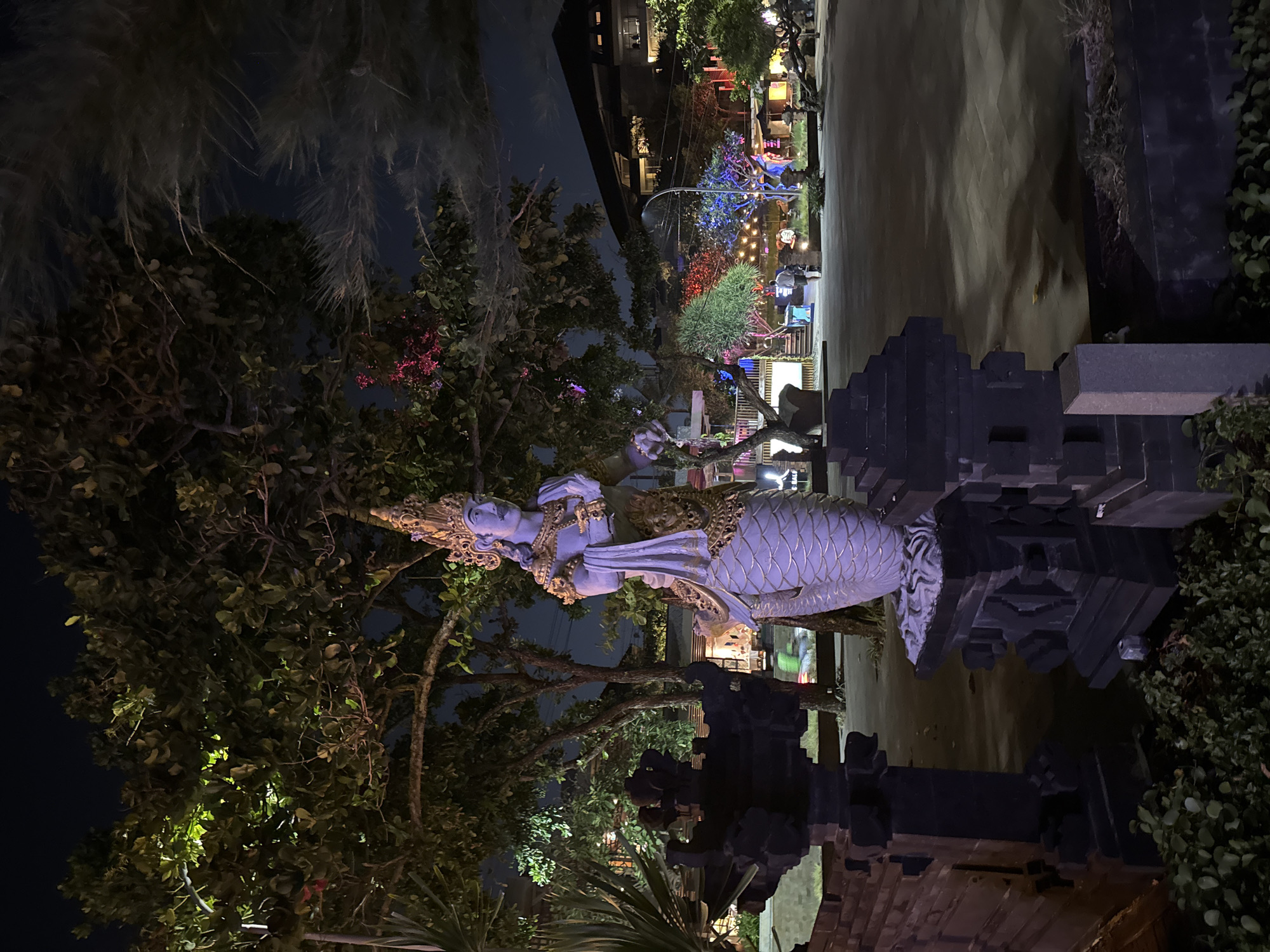 The image depicts a statue of a mythical creature, possibly a naga or a similar serpentine deity, in the foreground. The statue is illuminated, showcasing its detailed features, including scales and ornate headwear. It stands on a multi-tiered base, flanked by dark, stone architectural elements that resemble temple gates or pillars. In the background, a vibrant night scene unfolds in Kuta, Indonesia. Trees are illuminated with colored lights, creating a festive atmosphere. A building with multiple levels and lit windows is visible, suggesting a commercial or hospitality establishment. Several individuals are present in the background, some walking, others appearing to be seated or standing in groups, enjoying the ambiance. The overall environment suggests an outdoor plaza or promenade area, characteristic of a tourist destination in Kuta. The lighting is artificial, highlighting the contrast between the dark sky and the illuminated subjects and surroundings.