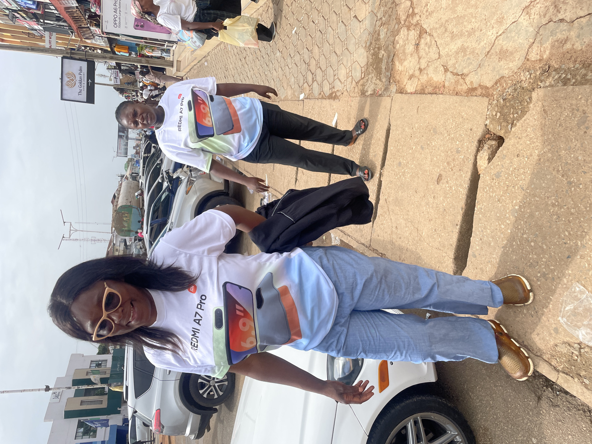 The image captures two individuals standing in what appears to be a street in Jos, Nigeria. Both are wearing white T-shirts with a graphic of the "REDMI A7 Pro" phone printed on them. One individual is wearing blue jeans and brown sneakers, while the other is wearing black pants and sandals.
The location is identified by a sign visible in the background that reads "The Golden Palm CAFE & JUICERY," along with other commercial signage such as "OPPO A6 Pro." Several parked cars line the street, including what seems to be a white SUV in the foreground and other vehicles in the background. The environment suggests an urban setting, with buildings and signs indicating commercial activity. The road and sidewalk surface are visible, with some areas showing signs of wear and disrepair.