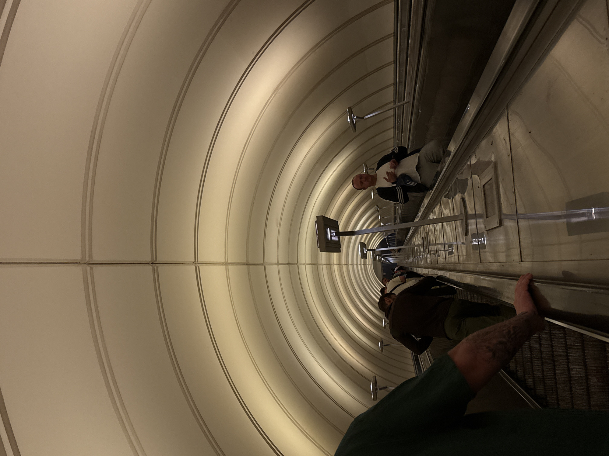 The image shows people on an escalator in a Moscow Metro station. The escalator appears to be going upwards and is enclosed within a long, arched tunnel that is illuminated by recessed lights. Several people are visible on the escalator, some facing forward and others facing backward. The ceiling of the tunnel is a light color with a ribbed design. The escalator handrails are metallic and reflective.