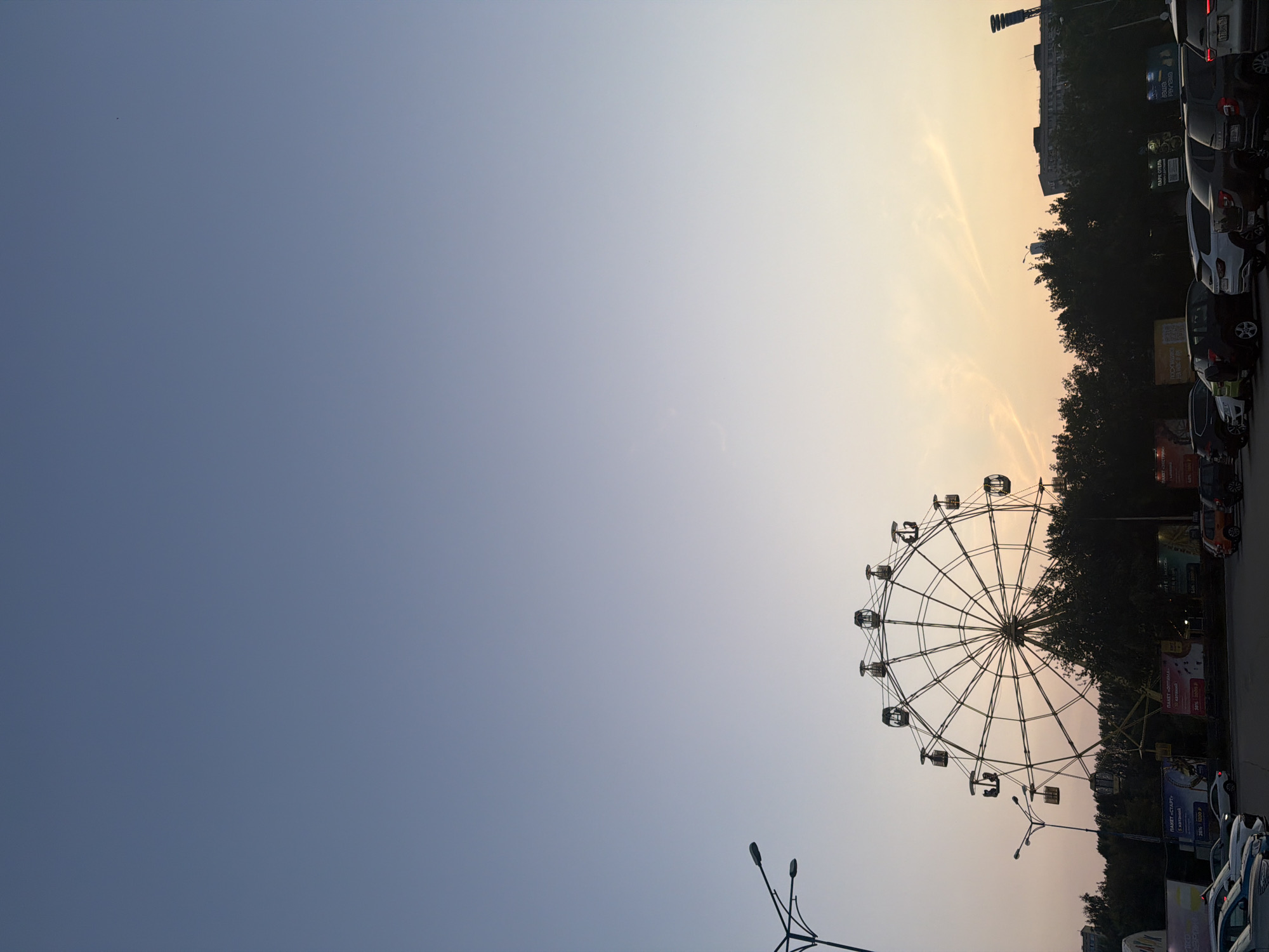 This image depicts an evening scene in the city of Tolyatti, Russia, likely at an amusement park or entertainment area adjacent to a parking lot. Dominating the frame is a large Ferris wheel, silhouetted against a gradient sky that transitions from a clear, pale blue at the top to a soft orange and yellow hue near the horizon, indicating sunset. Several gondolas are visible on the Ferris wheel, with some appearing to be occupied by people.
In the foreground, a row of parked cars is visible, suggesting this is a public space accessible by vehicle. The cars are of various makes and models, and their headlights are not illuminated, consistent with the fading daylight. Behind the Ferris wheel and cars, a dense line of dark green trees forms a backdrop. Above the trees and to the left, a street lamp with multiple lights is partially visible. In the distance, buildings can be seen, including what appears to be a tall structure with a spotlight or antenna. The overall atmosphere is calm and serene, with the natural light of the sunset casting a warm glow on the scene. The presence of advertisements on banners behind the Ferris wheel suggests a commercial or entertainment venue.