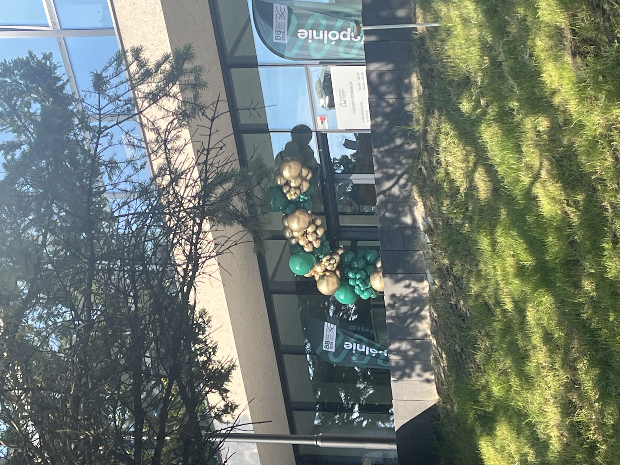 The image shows the entrance to a building with a large window and a small tree in the foreground. A balloon arch made of green and gold balloons is placed over the entrance. A flag with a logo and the word "spolnie" is also visible. The building appears to be made of concrete and glass. The ground in front of the building is covered with grass.