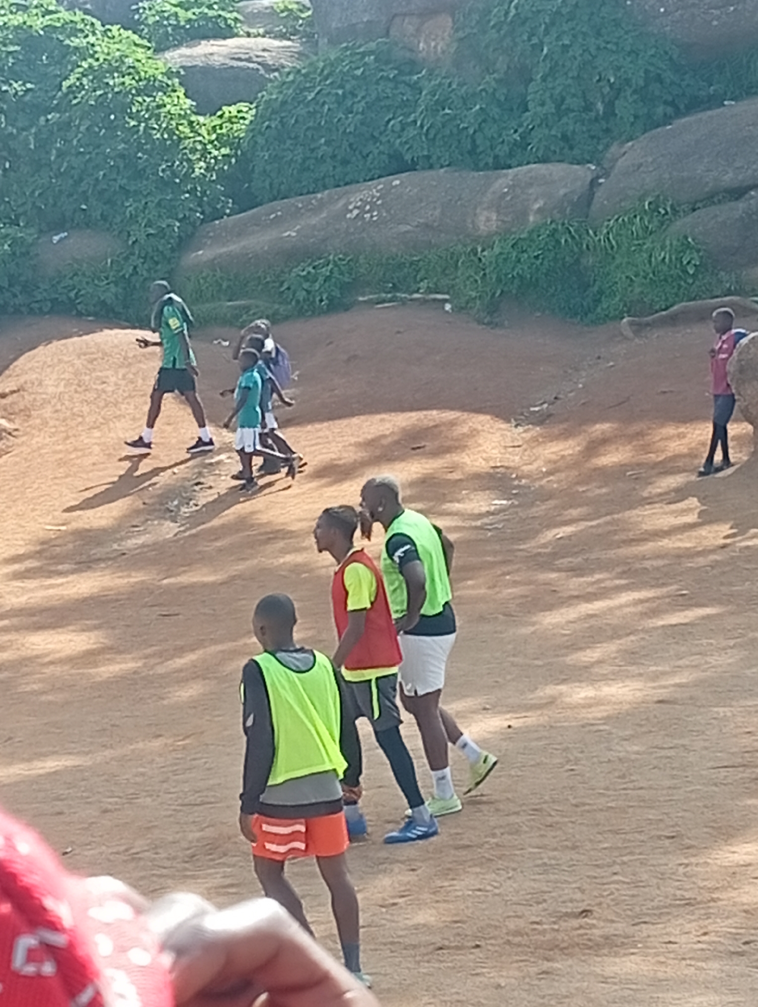 In the city of Jos, Nigeria, a group of children and young adults are engaged in a soccer training session on a dusty, uneven field. The environment is characterized by large, weathered boulders and lush green vegetation, suggesting a natural, undeveloped area. The participants are wearing athletic attire, including brightly colored vests, shorts, and sneakers, indicating their involvement in sports. Some are actively playing, while others appear to be observing or waiting for their turn. The lighting suggests it is daytime, with shadows cast by the individuals and the surrounding terrain.