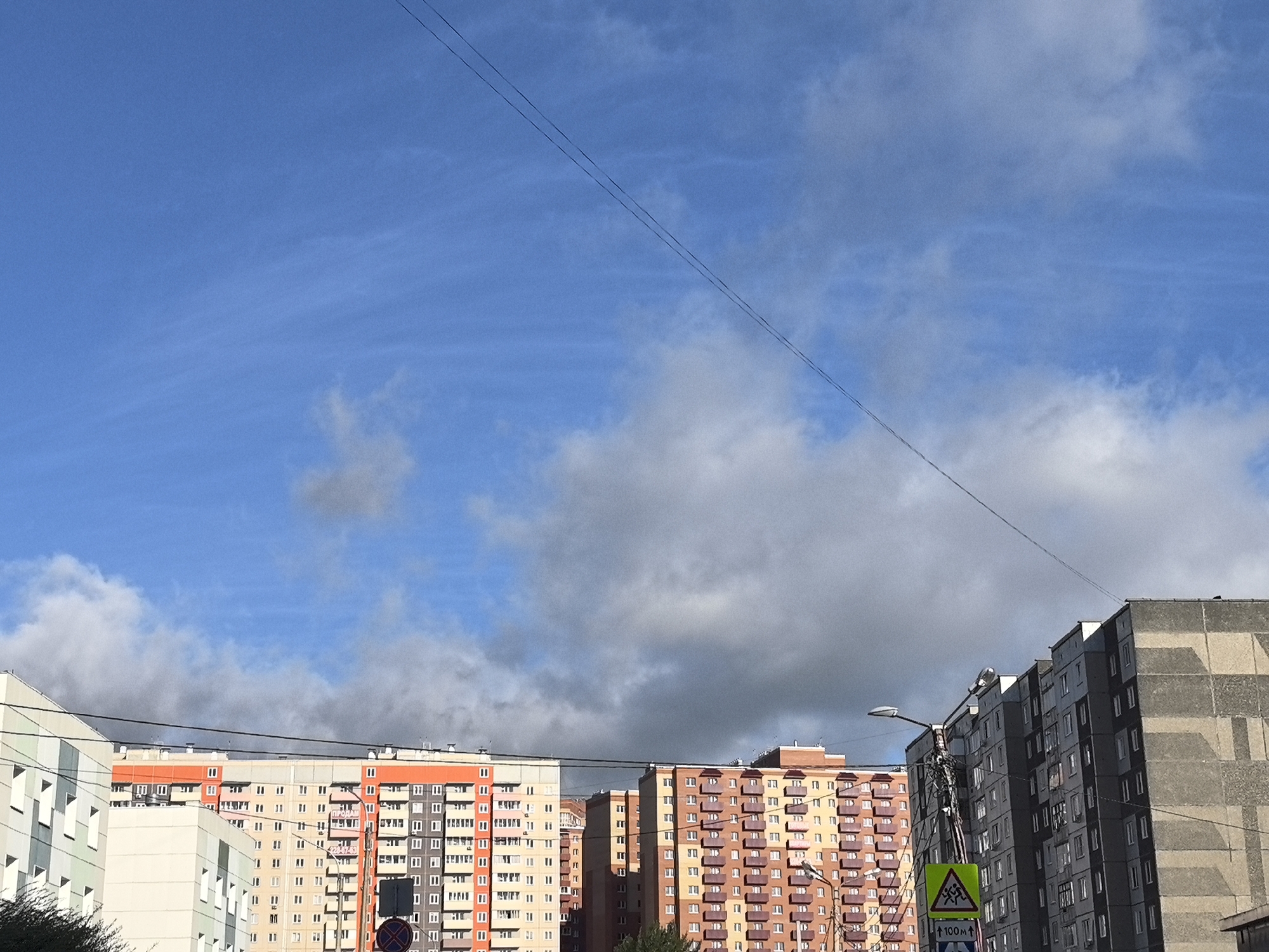 In the city of Unknown, Unknown, the image captures a view of multi-story apartment buildings under a partly cloudy blue sky. Two parallel dark wires traverse the sky diagonally from the upper left to the lower right. The buildings, constructed from concrete and exhibiting various colors and architectural styles, range from light gray and white to vibrant orange and pink. Some buildings feature balconies and numerous windows. A street light is visible on the right side, and a yellow traffic sign with a depiction of children and an arrow indicating 100 meters is mounted on a pole. Some sparse green foliage can be seen at the bottom left and right of the frame. The scene is an outdoor, daytime setting.
