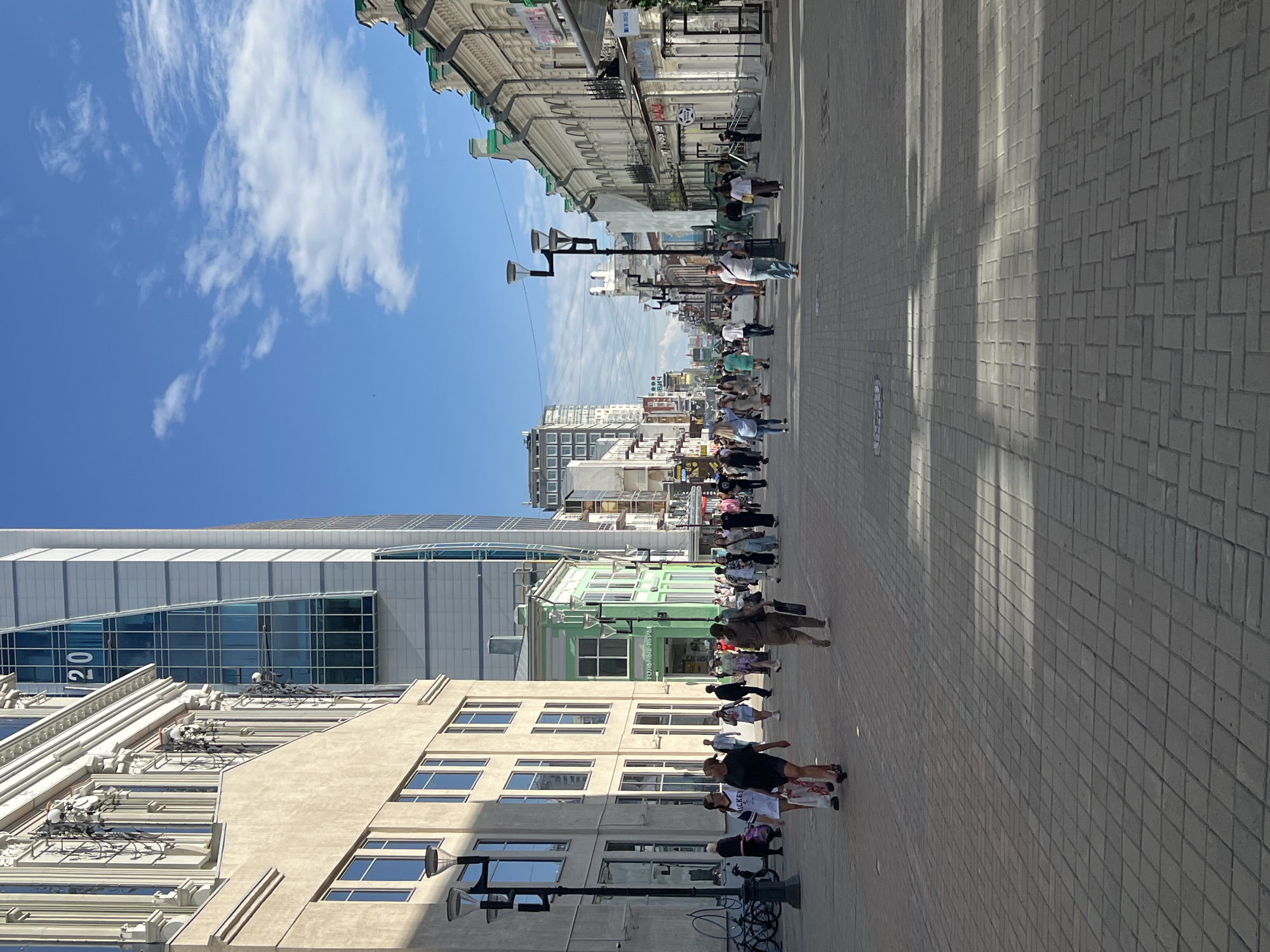 The image depicts a street scene in Yekaterinburg, Russia, on a sunny day with scattered clouds. A long line of people is walking down a pedestrianized street lined with buildings. The buildings vary in architectural style, from older, ornate structures to modern glass and steel constructions. Some buildings have green and cream facades, while others are a neutral beige or white. The street is paved with rectangular bricks, and long shadows indicate it's likely midday.
The people are dressed in casual summer clothing, suggesting warm weather. They are moving in a single file line, some walking in pairs or small groups. Their expressions are not clearly discernible due to the distance and resolution, but the overall atmosphere appears to be that of a leisurely stroll or perhaps participation in an organized event, given the orderly line.
Notable details include a modern skyscraper on the left with the number "20" visible on its glass facade. Street lamps with multiple bulbs are spaced along the street. There is some visible text on storefronts, but it is not clearly legible. The blue sky and bright sunlight are prominent features. The scene is a dynamic capture of urban life on a pleasant day in a Russian city.