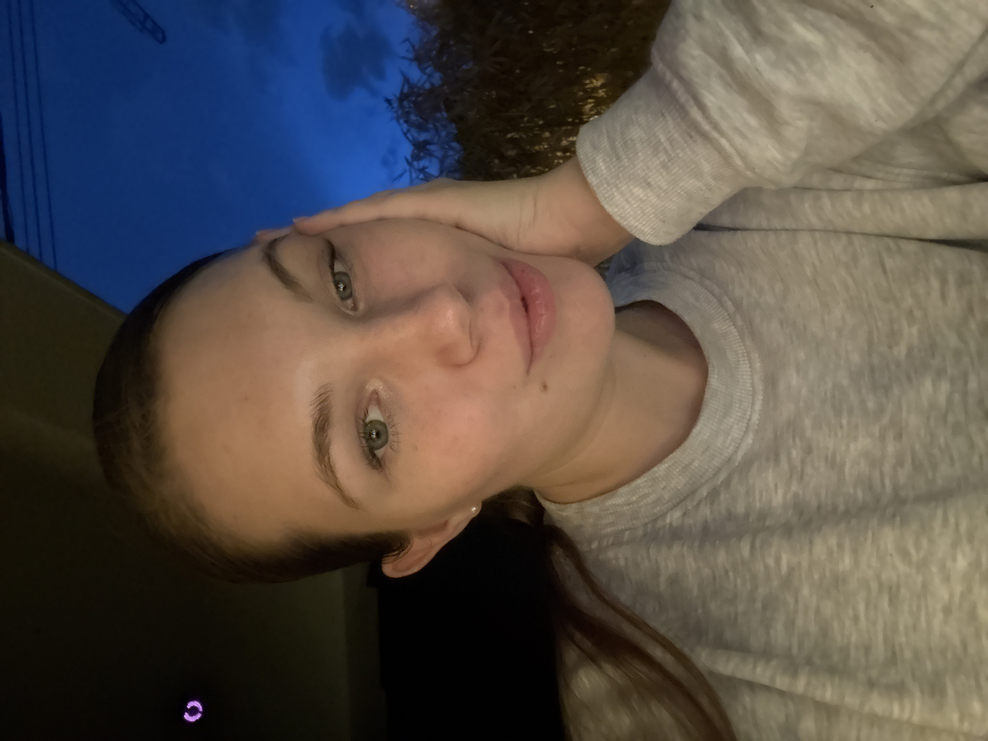 A portrait-style photograph of a light-skinned person with green eyes and light-brown hair is presented in a sideways orientation. The person is wearing a light grey sweatshirt and is touching their forehead with their right hand. The background is a mix of sky, vegetation, and a structure with visible power lines. There is no direct indication within the image that specifies a location in Belgrade, Serbia. No further details can be discerned without additional context.