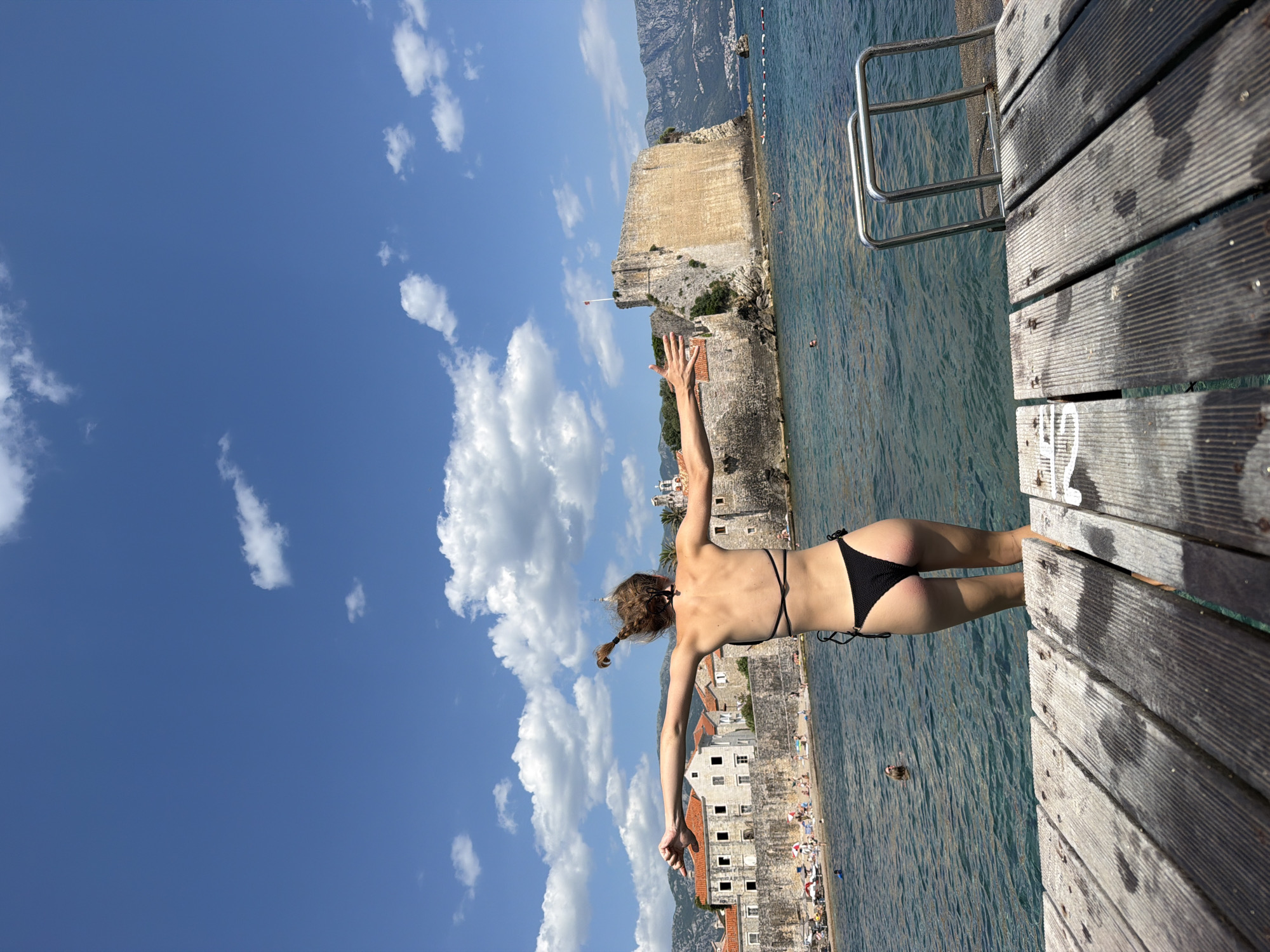 This media file captures a vibrant scene under a bright blue sky dotted with fluffy white clouds, suggesting a sunny day. The primary subject is a young woman in a black bikini poised on the edge of a wooden pier, her arms outstretched as if about to dive into the clear blue water. She has her hair in a braid that extends to her left. The pier itself is made of weathered wooden planks, with a metal ladder for entering and exiting the water.
In the background, a historical stone fortress with a weathered facade stands prominently against the coastline, a clear indicator of the location's rich history. Beyond the fortress, a cluster of old stone buildings with red-tiled roofs can be seen, leading to a hilly, mountainous landscape in the far distance. The sea is calm, with a few other swimmers visible in the distance, and a beach area with sunbathers is partially visible along the shore.
The overall mood is one of summer joy and adventurous spirit. The bright sunlight and the woman's dynamic pose evoke a feeling of freedom and exhilaration. The image seems to have been taken in the afternoon, given the strong shadows cast by the pier. While no explicit text is visible, the architectural style of the buildings and the fortress strongly suggest a location in Southern Europe, specifically Budva, Montenegro, as indicated by the provided context. The perspective is from a low angle, looking upwards, which emphasizes the vastness of the sky and the height from which the woman is about to jump.