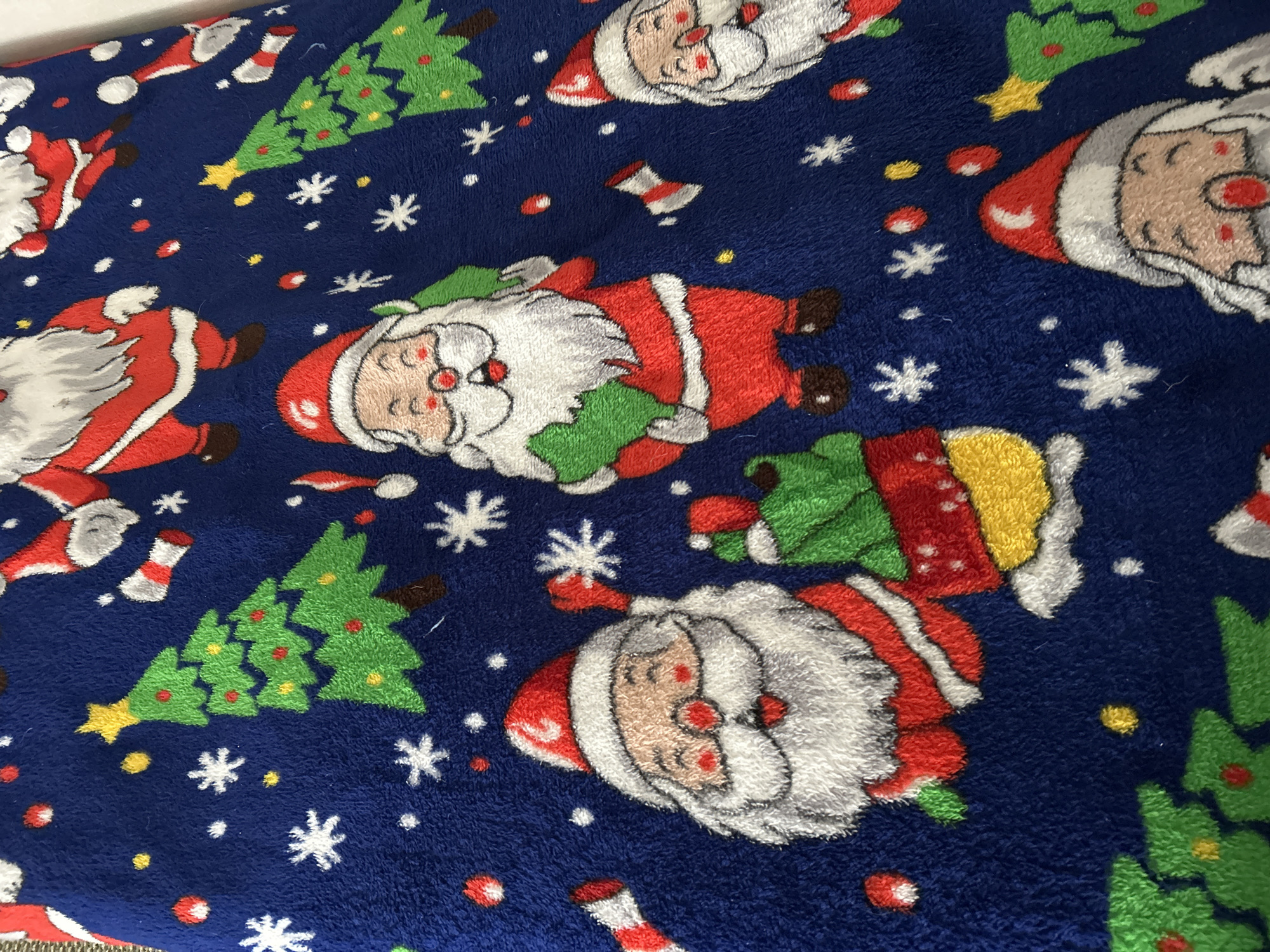 The image shows a close-up of a blue fabric with a repeated pattern of Christmas-themed elements. The pattern includes illustrations of Santa Claus, Christmas trees decorated with small balls, white snowflake shapes, and small red and yellow circles. Some striped socks are also present. The Santa illustrations vary in their pose. The location is not evident from the image.