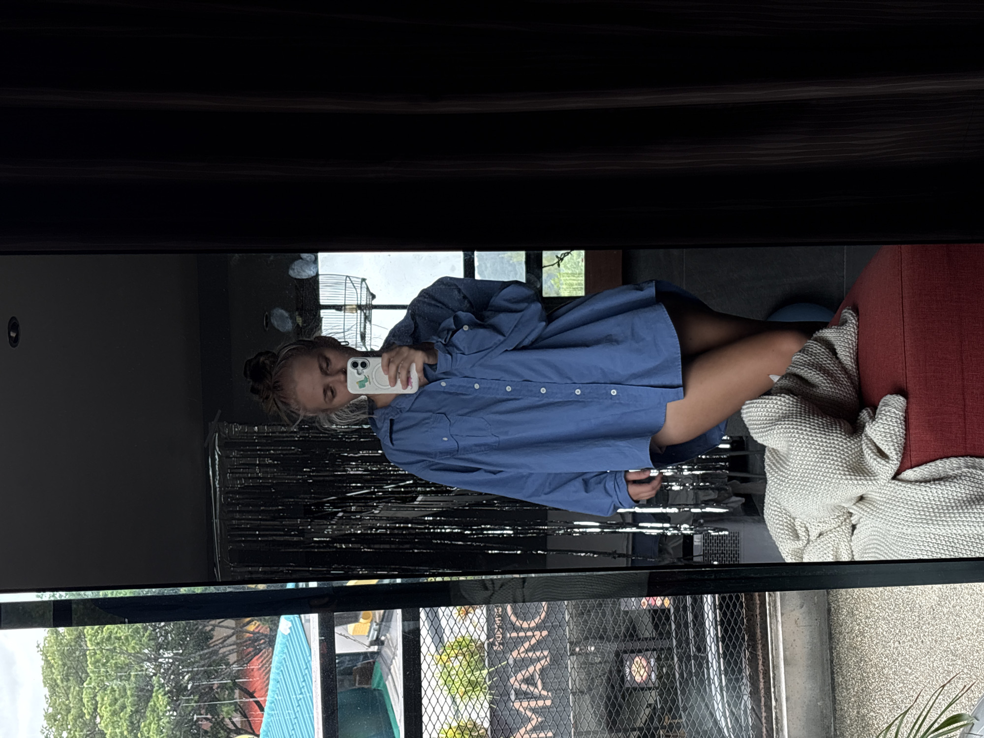 A young woman with blonde hair in a bun is reflected in a full-length mirror, taking a selfie with a white smartphone. She is wearing an oversized, light blue button-down shirt, which is open and trails down her thighs, revealing her legs. Her left arm is bent, holding the phone up, and her right hand rests on her hip. Behind her, in the reflection, are silver tinsel curtains, a birdcage, and a view of trees and a building with a blue roof. In the foreground of the reflection, a beige knitted blanket is draped over a red ottoman. The scene appears to be indoors, possibly a residential setting, given the presence of the ottoman and blanket, and the view outside suggests a semi-urban or tropical environment. The overall lighting is natural, coming from the window.