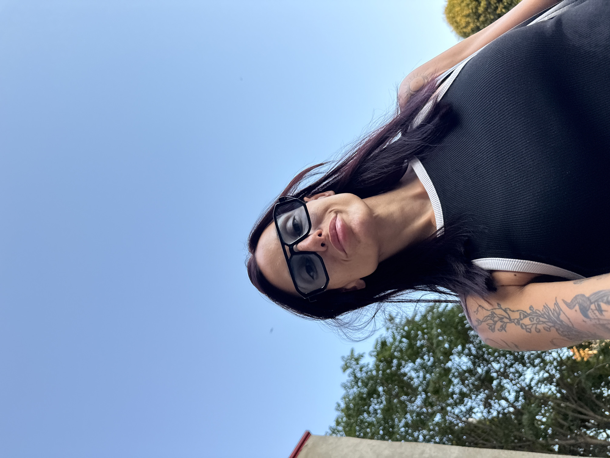 A low-angle, waist-up shot shows a woman with dark hair and sunglasses posing outdoors. She is wearing a black tank top with white trim and has a tattoo on her left arm. She is looking towards the camera with a slight smile. In the background, a tree and part of a building are visible against a clear blue sky. The image is an outdoor portrait taken in Pleven, Bulgaria.
