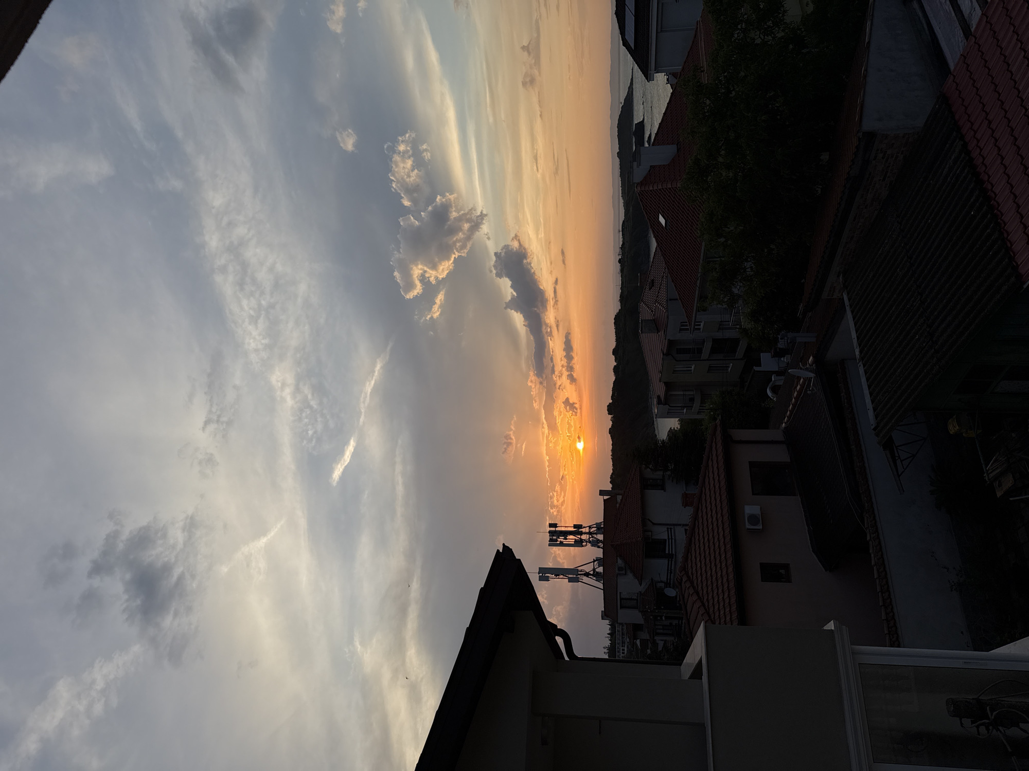 The media file presents a panoramic landscape view of a coastal or lakeside town, identified as Chernomorets, Bulgaria, during sunset. The image is captured from a high vantage point, likely a balcony or rooftop, looking towards the horizon where the sun is setting.
The sky dominates the upper portion of the frame, showcasing a beautiful blend of colors. On the left, the sky is a pale blue and grey with wispy, streaky clouds. Towards the right, it transitions into warm hues of orange and yellow, intensified by the low-hanging sun. The sun itself is visible as a bright, golden orb near the horizon, partially obscured by fluffy clouds that glow with the golden light. Some darker, more defined clouds are scattered across the mid-sky. A small bird can be seen in flight against the lighter sky on the left side of the sun.
Below the colorful sky, a residential area unfolds with numerous buildings, many featuring distinctive red tile roofs. These structures appear to be multi-story houses or apartments, nestled among green trees and foliage. Several communication or cellular antenna towers stand tall in the mid-ground, silhouetted against the bright sky. In the far distance, a calm body of water, likely the Black Sea, meets a hazy, forested landmass on the horizon.
The immediate foreground is characterized by the dark silhouette of a rooftop with a visible gutter, and parts of a white, modern-looking balcony or terrace structure. This foreground element is largely in shadow, contrasting with the illuminated sky and distant features. The overall mood is serene and picturesque, capturing the tranquil beauty of dusk over a coastal town. No discernible human activity or text is visible in the image.