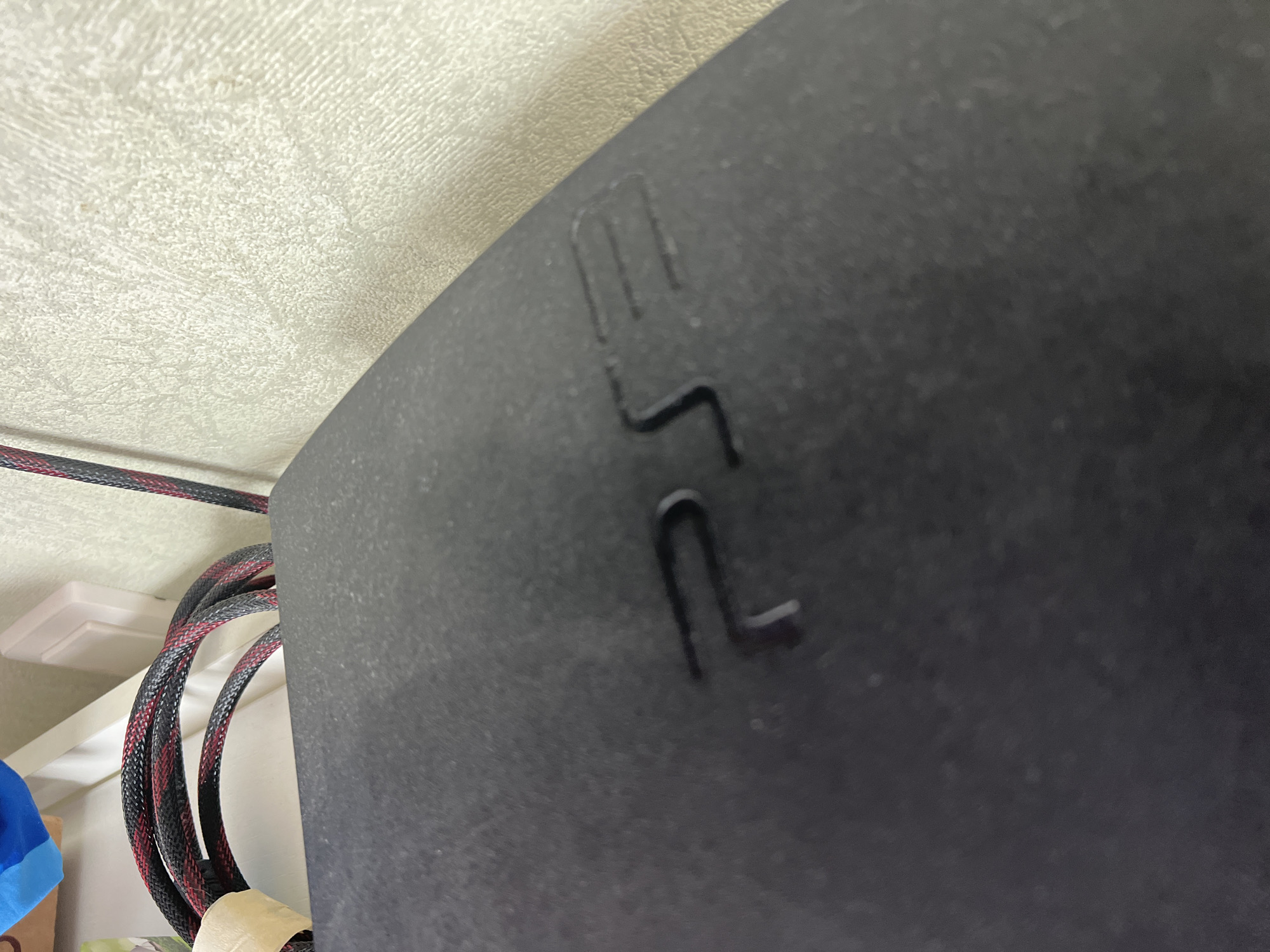 The image shows a close-up of the black casing of a PlayStation 3 console, with the "PS3" logo embossed on it. The console is resting on a textured, light-colored surface, possibly a wall. In the background, a bundle of black and red cables is visible, wrapped around something out of frame. There are no people or any identifiable elements of the city of Sochi, Russia, in this image.