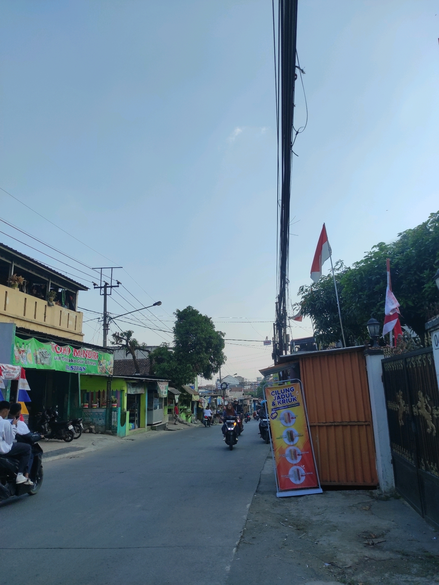 The image depicts a street scene in Padalarang, Indonesia, under a clear blue sky. The street is paved and appears to be a commercial area with several small shops lining the right side. On the left, a two-story building with a yellow facade has balconies where people are visible. A green banner with text and images hangs in front of a shop. Several motorcycles are parked in front of the shops and some are in motion on the street. Two young men are riding a motorcycle in the foreground, heading away from the camera. A tall pole with numerous black cables runs vertically through the center of the image. Two Indonesian flags are displayed on poles near the right side of the street, one larger than the other. A tall, orange, corrugated metal door is visible behind the flags. A standing sign with yellow and red colors is placed on the sidewalk in front of the orange door, advertising "CILUNG JADUL & KRIUK" with images of food items. The environment suggests a warm day in a bustling urban or semi-urban setting.