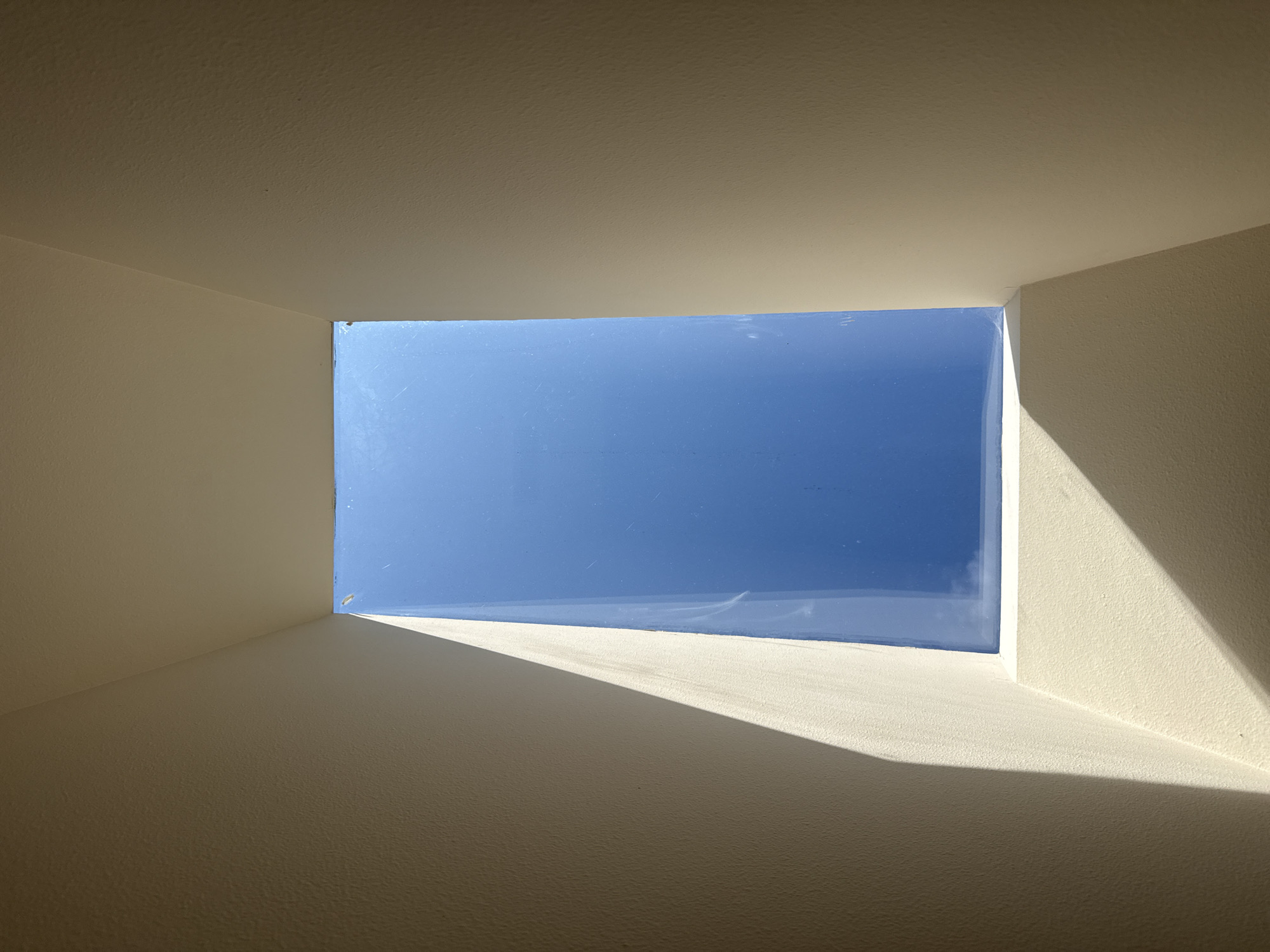 An outdoor, high-angle, medium shot shows a rectangular skylight against a bright blue sky. The skylight is framed by cream-colored, stucco-textured walls that converge inwards at the top. Sunlight casts a sharp, triangular shadow across the bottom left portion of the frame, obscuring part of the wall and the bottom edge of the skylight. The sky visible through the skylight is clear and a uniform shade of blue, with no visible clouds or celestial bodies. The image appears to be taken from within a building looking upwards through the skylight. The specific location in Sammamish, United States of America, is not determinable from the image.