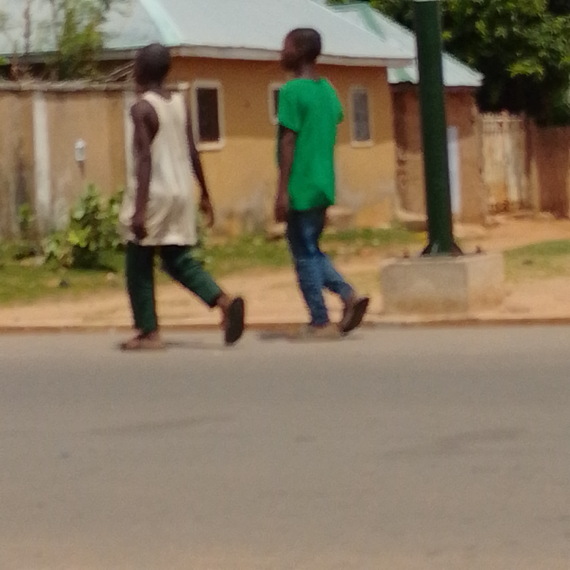 Two individuals, one wearing a white sleeveless top and green trousers, and the other a green t-shirt and blue jeans, are walking on a paved street in Gombe, Nigeria. The person in the white top is slightly ahead and to the left of the person in the green t-shirt. They are walking in the same direction. In the background, there is a light-colored building with windows, a fence, and some greenery. A dark green pole is also visible on the right side of the image. The overall scene suggests a casual outdoor environment in a residential or commercial area of Gombe.
