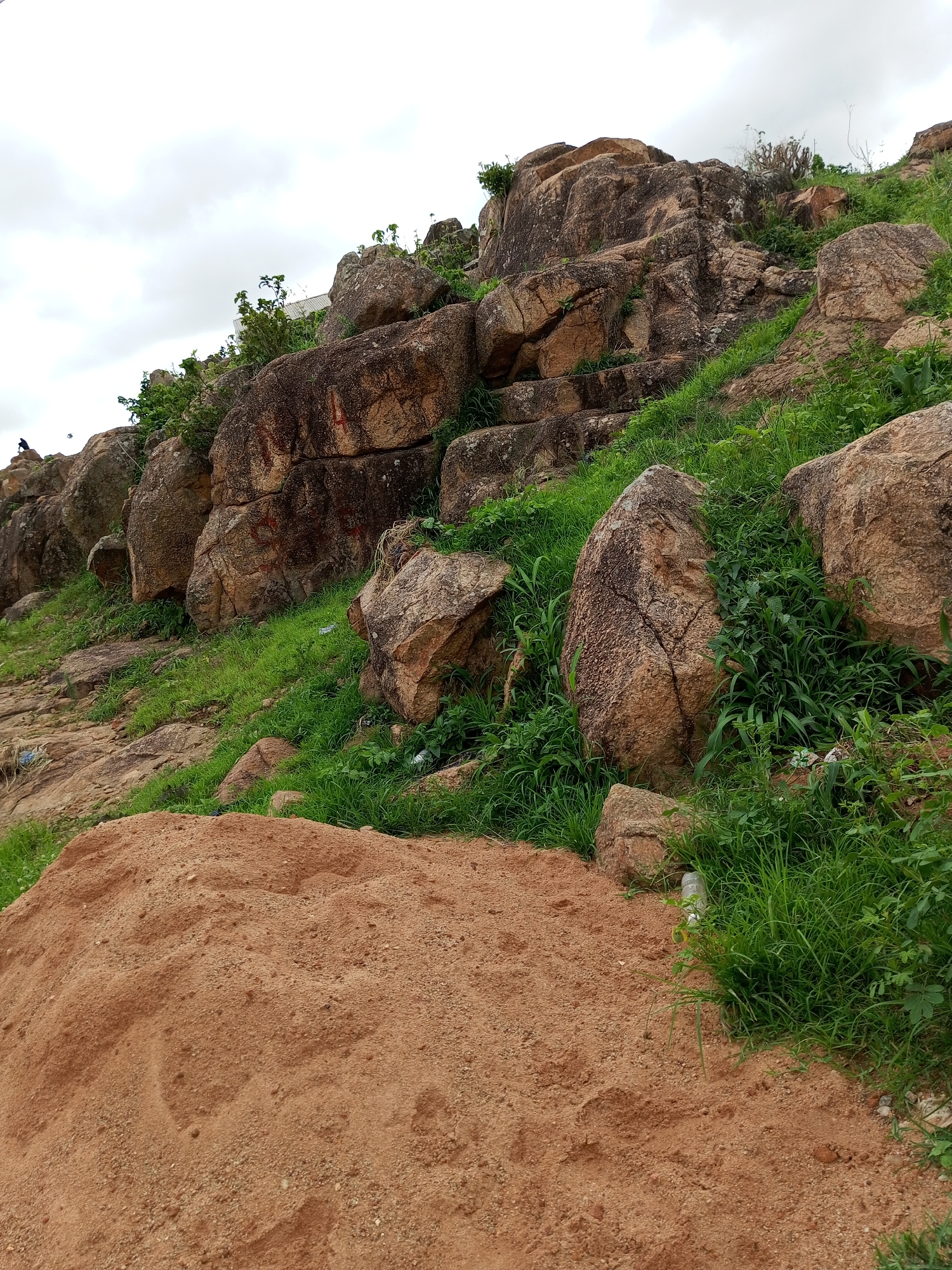 The image depicts a rocky, overgrown hillside under a cloudy sky. A large pile of reddish-brown sand dominates the foreground. The hillside is covered in sparse green grass and interspersed with large, weathered rocks. Some of the rocks show signs of erosion and cracking, with reddish-brown markings on their surfaces. In the middle ground, a small, dark figure, possibly a person, is visible on a rock in the upper left portion of the image. The environment appears to be natural and somewhat undeveloped, with no clear signs of a constructed path or significant human activity beyond the pile of sand and the distant figure. There is no indication of the specific location being within the city of Gura Top, Nigeria.