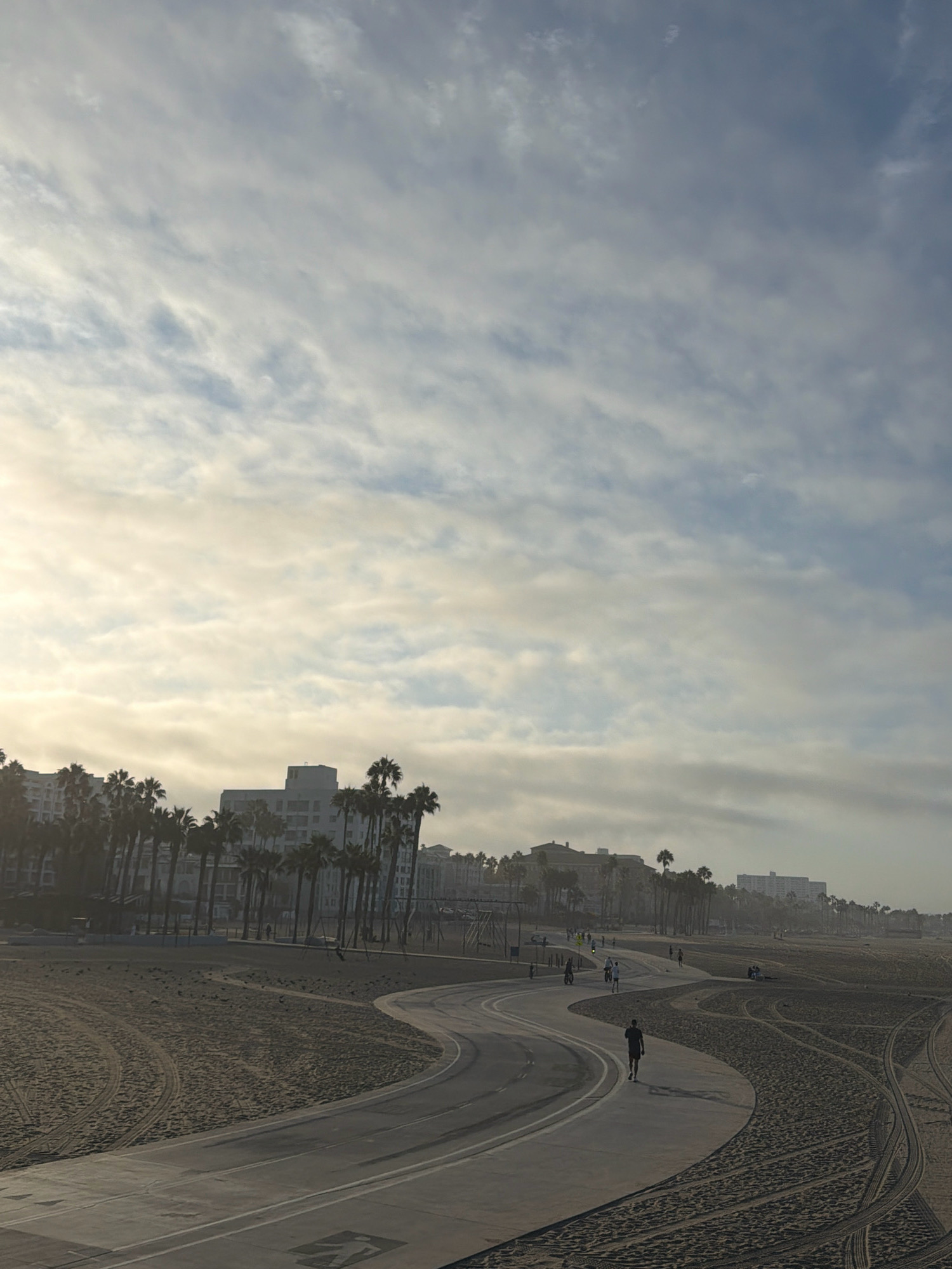A paved pathway curves through a sandy beach, bordered by palm trees and buildings in the distance. The sky is filled with a mix of clouds and sunlight. A lone figure walks along the pathway, casting a shadow on the ground. Several other individuals are visible in the background, engaged in activities such as walking and cycling. The beach has tire tracks from vehicles or bicycles on the sand. The architecture of the buildings suggests a coastal urban environment.