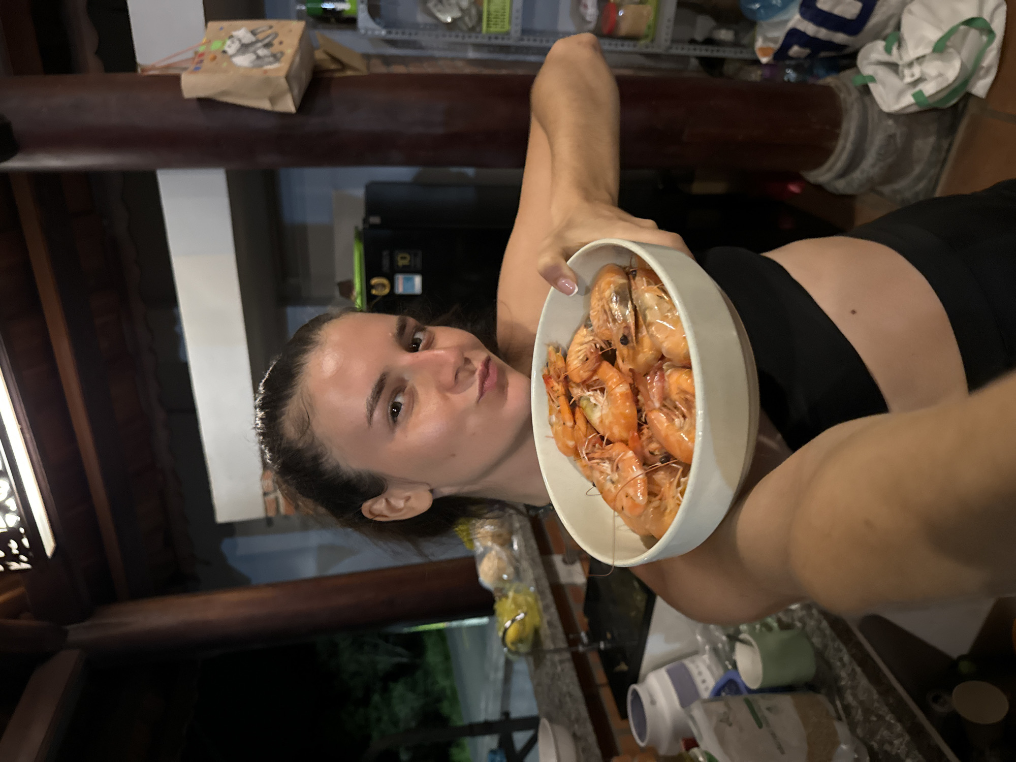 The image depicts an outdoor or semi-enclosed kitchen and dining area in Mũi Né, Vietnam, illuminated by artificial lighting at night. A young adult female is positioned centrally, holding a white ceramic bowl filled with numerous cooked, orange-red shrimp towards the viewer with both hands. She has dark hair pulled back and is wearing a black crop top and black lower garment, revealing a small mole on her abdomen. Her lips are pursed, and she maintains direct eye contact with the camera.
To her left, a light-colored, possibly granite or stone, countertop is visible, featuring a sink with a faucet, a large white container, a green mug, and various other bottles and packaged items. Beyond the counter, out of focus, glimpses of outdoor greenery and distant artificial lights suggest an exterior setting. To the right, a prominent dark wooden pillar supports the structure, with a brown paper bag displaying a panda design hanging from it. In the background, a black refrigerator with a "10" sticker and a partially visible metal shelving unit stocked with various items like jars are present. The ceiling above consists of dark wooden beams, with a decorative light fixture also visible. A white and blue bag is situated on the floor near the right pillar.