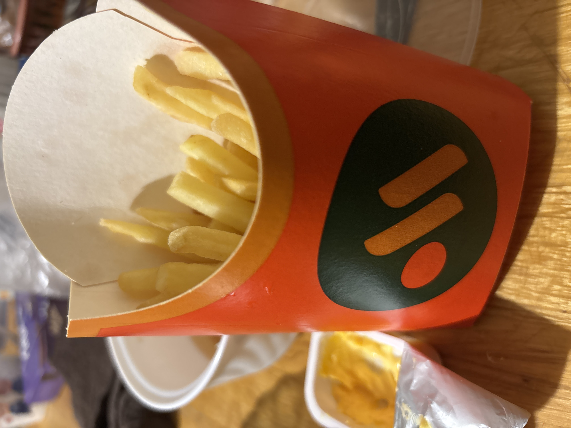 A close-up, slightly tilted, indoor shot displays a nearly full carton of french fries. The orange and green carton features a logo. The fries themselves are pale yellow. Below the carton are other food items, including containers. The setting appears to be on a wooden table, suggesting a home or restaurant environment. The location is presumed to be in Vsevolozhsk, Russia.