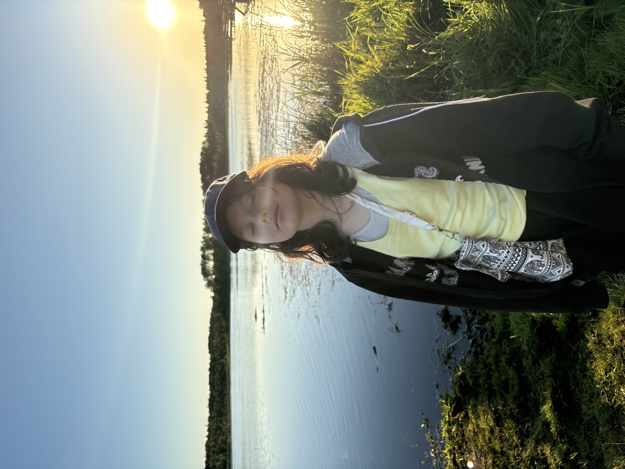A young person stands by a body of water, facing forward with their eyes closed, bathed in the golden light of a setting sun. They are wearing a dark baseball cap, a light yellow top layered under a dark zip-up hoodie, and black pants. A small, patterned crossbody bag is slung over their shoulder. The background features a calm lake reflecting the sky, with a treeline on the far shore and tall grasses in the foreground. A wooden dock is visible in the distance to the right. The scene is illuminated by the warm glow of the sun, creating long shadows and a serene atmosphere. The location is identified as being in Chikino, Russia.
