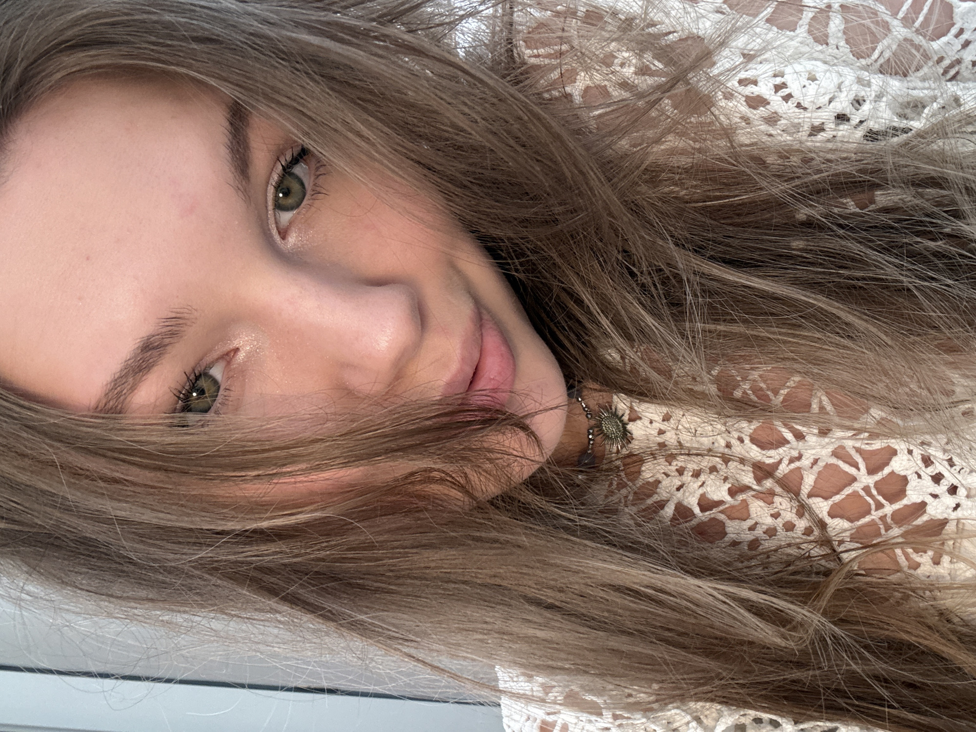 A close-up, angled shot of a Caucasian woman. She has long, light brown hair that partially obscures her face. Her skin appears fair. She has green eyes and a gentle smile. She is wearing a white, lace-patterned top. A necklace with a circular pendant is visible at her neck. The background is mostly out of focus, with a suggestion of a white surface at the bottom of the frame. There is no information to indicate the location is in Vozdovac Urban Municipality, Serbia.