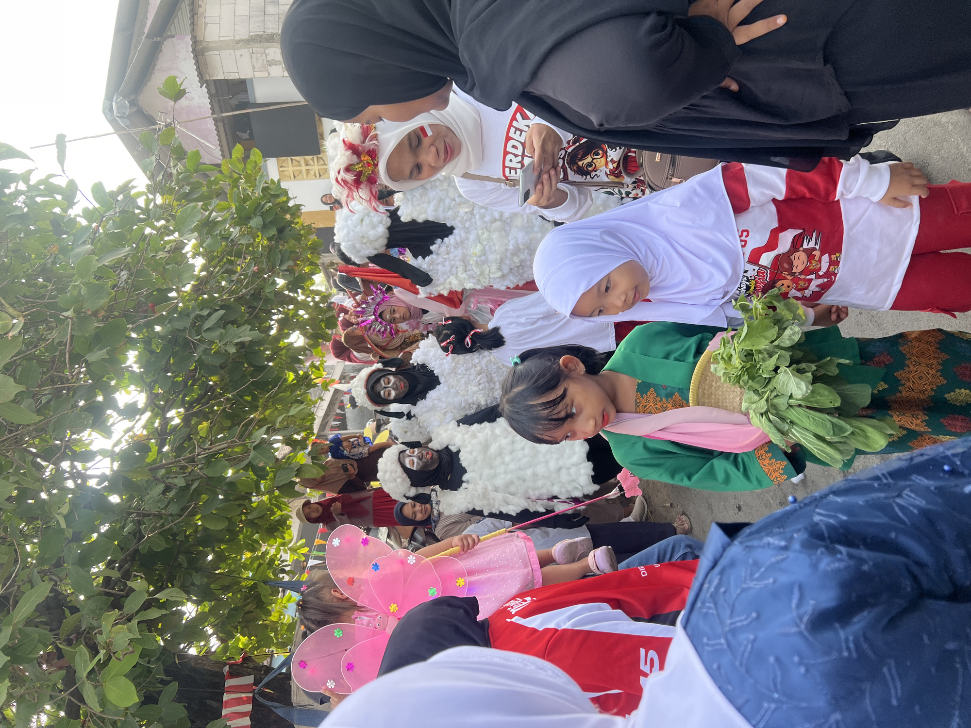 In the city of Klapanunggal, Indonesia, a group of children and adults are gathered outdoors, seemingly for a parade or festival. Several children are dressed in costumes. Two children wear white fuzzy costumes resembling sheep, complete with black face paint. Another child is dressed as a fairy with large pink wings. Some participants have facial paint and elaborate headwear. The crowd includes people of various ages, many of whom are wearing traditional Indonesian clothing, including hijabs. The environment appears to be a street scene with trees and buildings in the background. Red and white banners are visible, suggesting a patriotic celebration. The overall atmosphere suggests a lively community event.