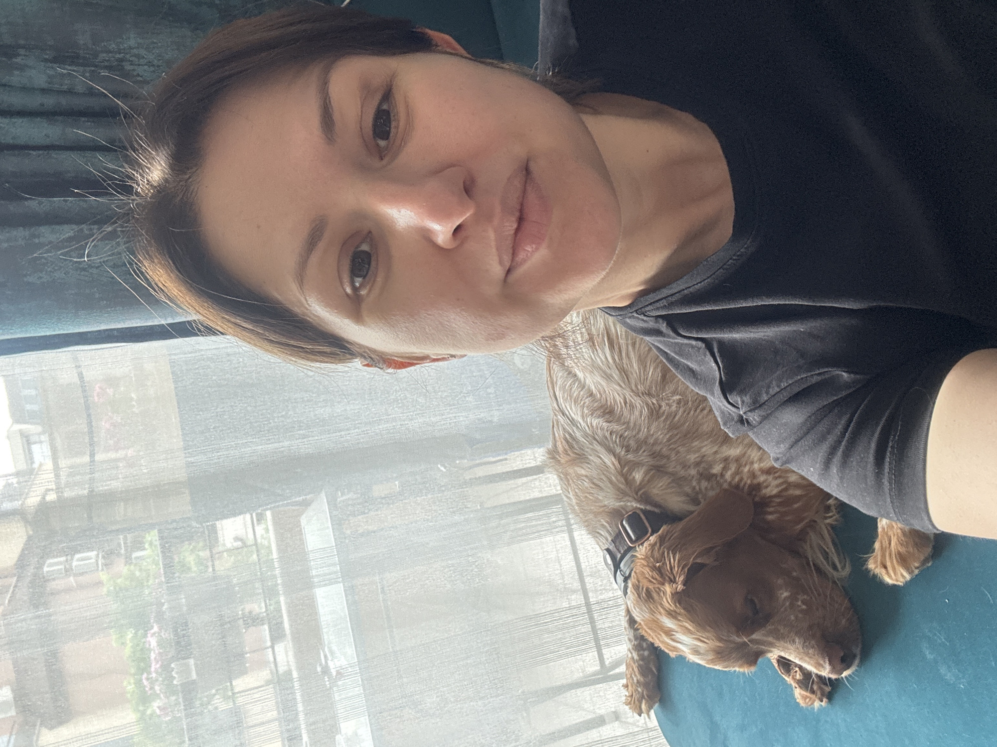 A woman and a dog are visible in the scene. The woman is fair-skinned with brown hair and is wearing a black t-shirt. Her face is centered in the frame, and she appears to be looking directly at the camera. A brown and white dog lies next to her, wearing a black collar. Behind them is a window covered with a sheer white curtain. Outside the window, buildings and greenery can be seen. Based on the style of architecture and greenery, the location may be in Sofia, Bulgaria. The photograph seems to be taken indoors in a well-lit setting.