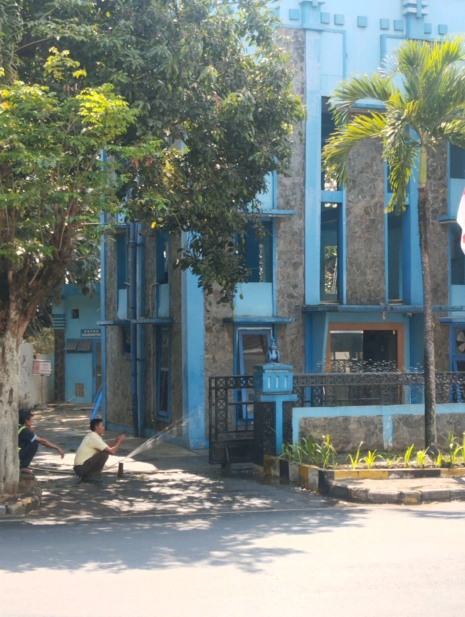In Malang, Indonesia, on a sunny day, two individuals are present in front of a blue and stone-clad building. One person, wearing a yellow shirt and dark pants, squats and holds a hose, directing a stream of water onto the ground. The other person, in a blue shirt and dark pants, sits on a curb to the left. The building features a distinctive blue facade with stone accents and multiple windows. A palm tree stands to the right of the building, and lush green trees are visible on the left. The foreground shows a paved area with tree shadows.
