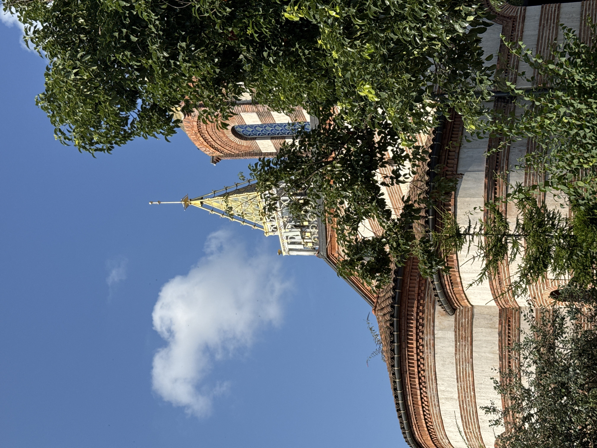 The image shows a view of the Holy Trinity Church in Batumi, Georgia, under a clear blue sky with a single white cloud. A large, leafy green tree partially obscures the facade of the church, which is constructed with alternating bands of red brick and white stone. The church features a tiled roof, arched windows, and a distinctive golden spire topped with a cross. The architectural style appears to be of Orthodox Christian design. No people are visible in the scene, and the environment is dry and sunny.