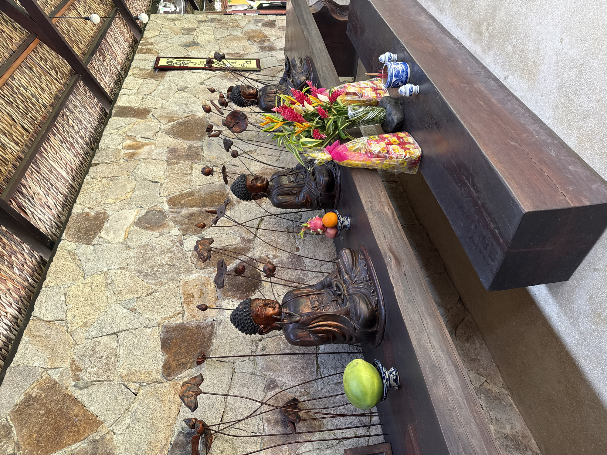 The image shows a Buddhist altar in Tay Nha Trang Ward, Vietnam. Four bronze Buddha statues are arranged on a dark wooden shelf against a stone wall. Each statue is accompanied by metal lotus plant decorations. Offerings are placed on the shelf, including fresh tropical flowers, packaged snacks, incense sticks in blue and white ceramic holders, a green mango, and small fruits in bowls. The ceiling is covered with a woven mat. The environment suggests a religious or spiritual space.