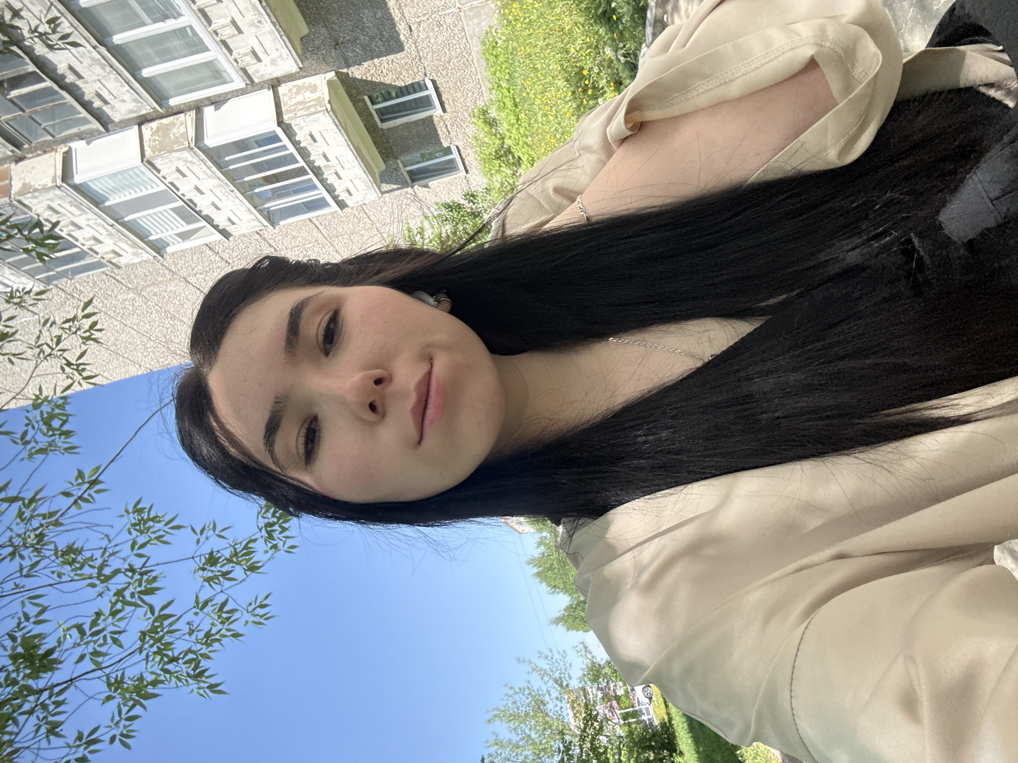 A young woman is taking a selfie outdoors. She has long, dark hair and is wearing a light-colored top. The background includes an apartment building and trees under a blue sky. Based on the architecture of the apartment building, specifically the window styles and overall design, the location is likely in Izhevsk, Russia.