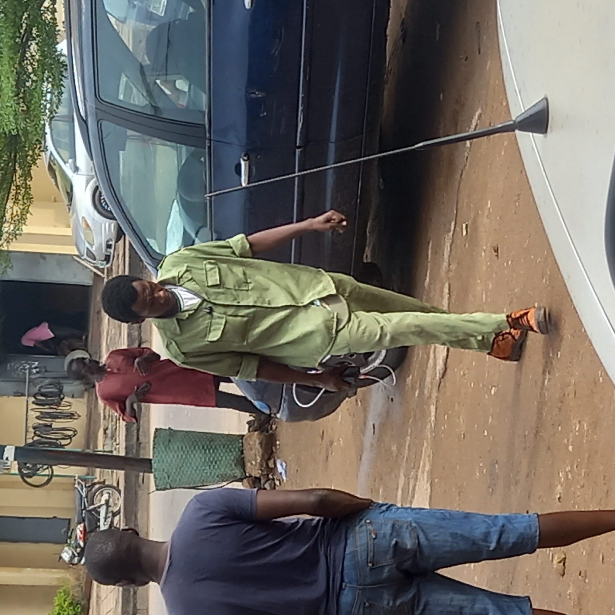In Sardauna, Nigeria, a man in a light green uniform stands on a paved street next to a dark blue car, holding a long, thin rod with a conical tip that touches the side of a white vehicle. He wears orange shoes and appears to be working on or inspecting something related to the car's tire. Another man in a red shirt stands nearby, and a man in a blue shirt and jean shorts is walking by. The background includes a building with bicycle parts hanging outside. A motorcycle and other vehicles are also visible. The environment suggests a roadside repair or inspection scene.