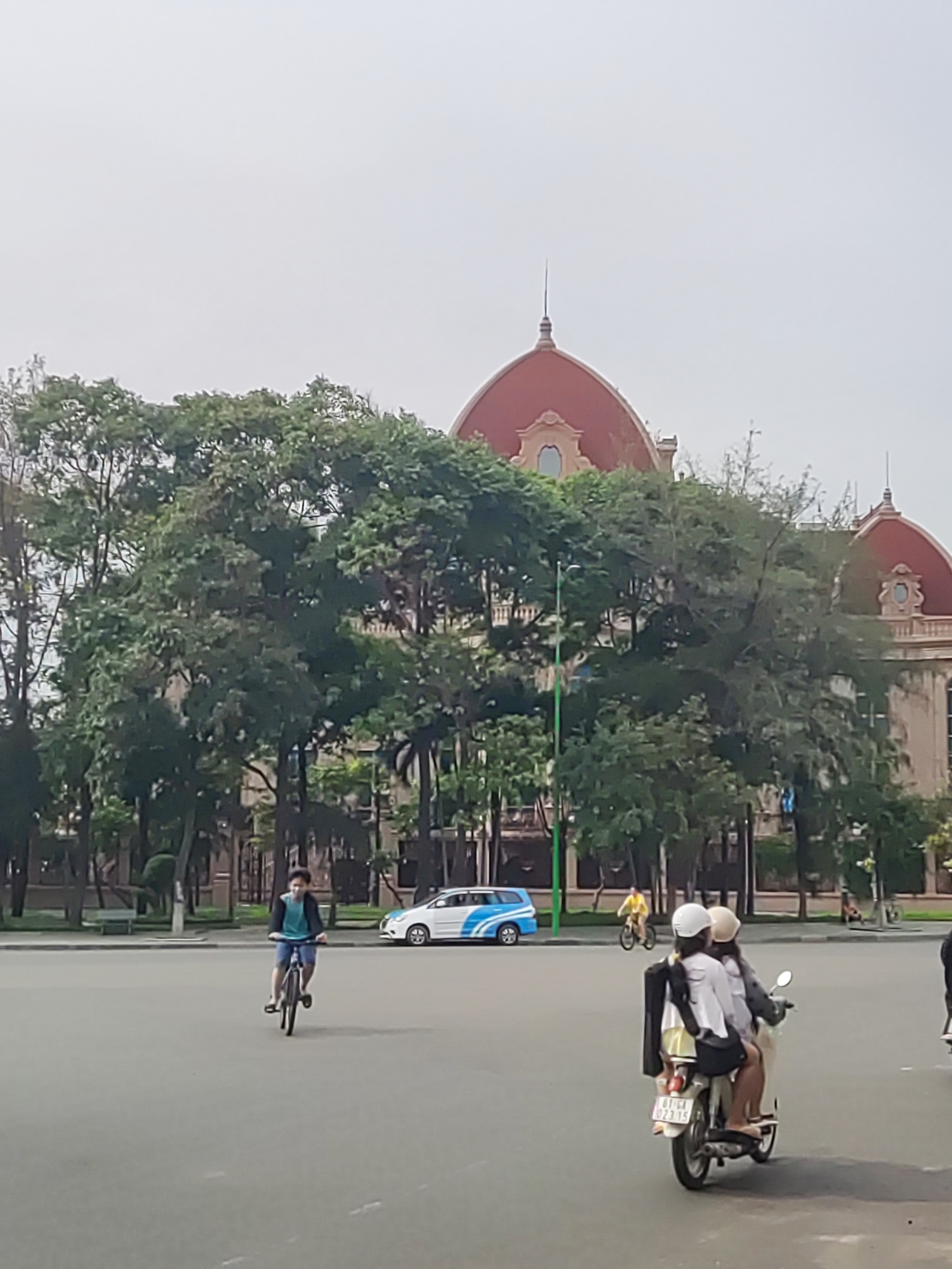 The image shows a large square in an urban environment. A large building with two reddish domes is partially obscured by trees in the background. A white car with a blue stripe is parked in the background. A man rides a bicycle on the left. Two people are riding a scooter on the right. Another person rides a bicycle in the background. A few other people are visible in the distance. The sky is overcast. The location appears to be in Unknown, Unknown, based on the license plate and architectural style.