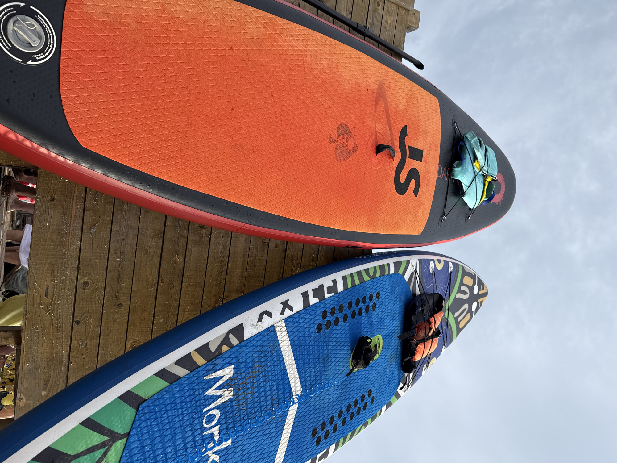 Two stand-up paddleboards are laid out on a wooden dock. The top board is orange with a black edge and red trim. It has a textured grip pad and black handles. A black paddle rests on top of it. A blue and yellow bag is attached to the board with bungee cords. The bottom board is blue with a colorful, abstract pattern. It also has a textured grip pad and black handles. A leash and a life vest are attached to this board. The paddleboards are positioned diagonally, with the sky visible in the background. The lighting suggests it is daytime, and the cloudy sky indicates potentially overcast weather. The wooden dock suggests an outdoor, waterfront location. The scene is static, with no activity taking place, but the presence of water sports equipment implies a setting where these activities are common. Text is visible on both boards. The top board has the logo "JS". The bottom board has the word "Morko" in large white letters and other smaller text that is illegible.