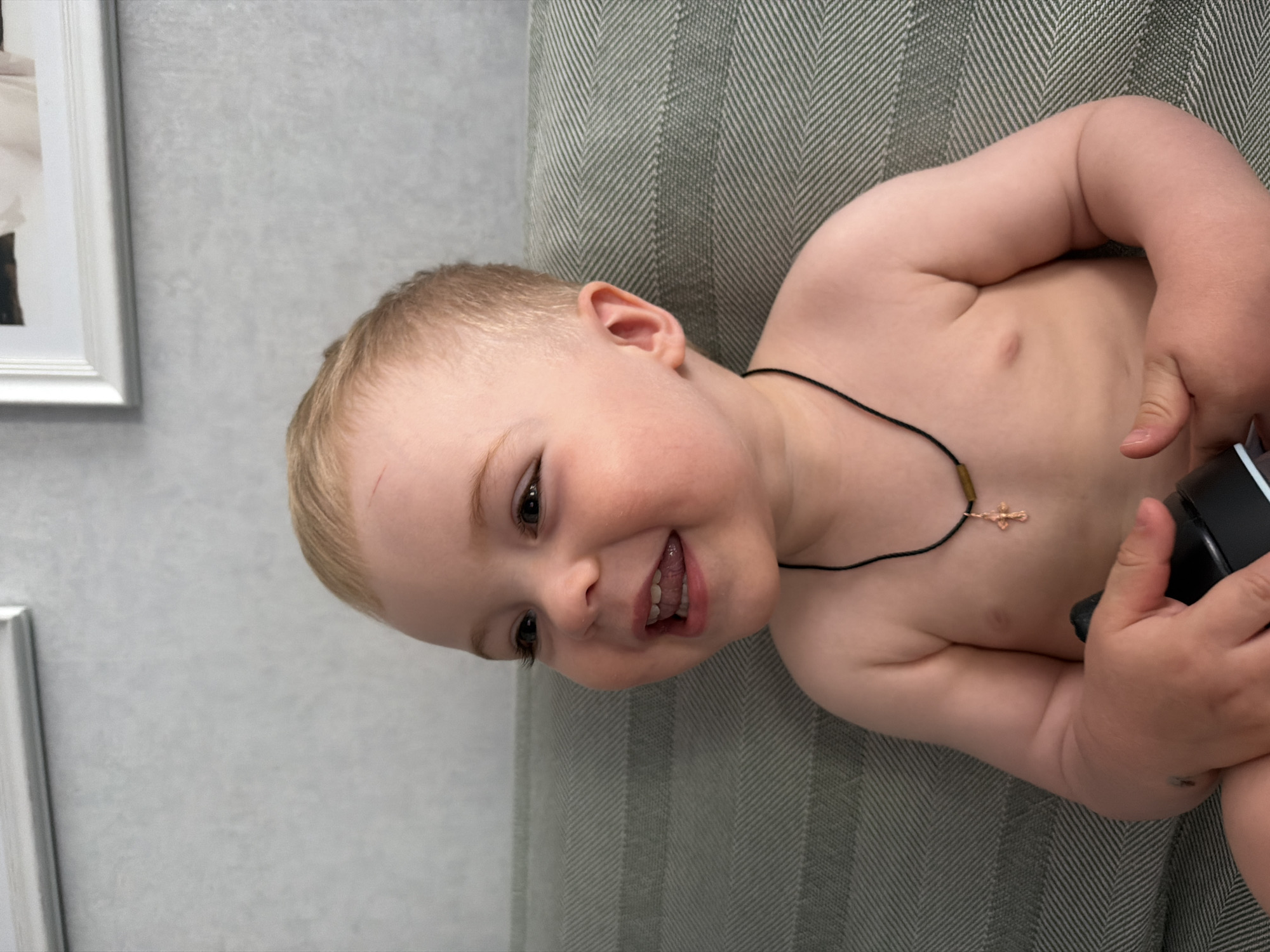 A young, fair-skinned child lies on a gray, vertically-striped upholstered surface. The child is shirtless and wears a thin black cord necklace with a small gold cross. The child smiles, exposing teeth. The child holds a black object in their right hand. A framed picture hangs on a gray wall to the left of the child. The location is unknown, and there is no evidence it is in Votkinsk, Russia.