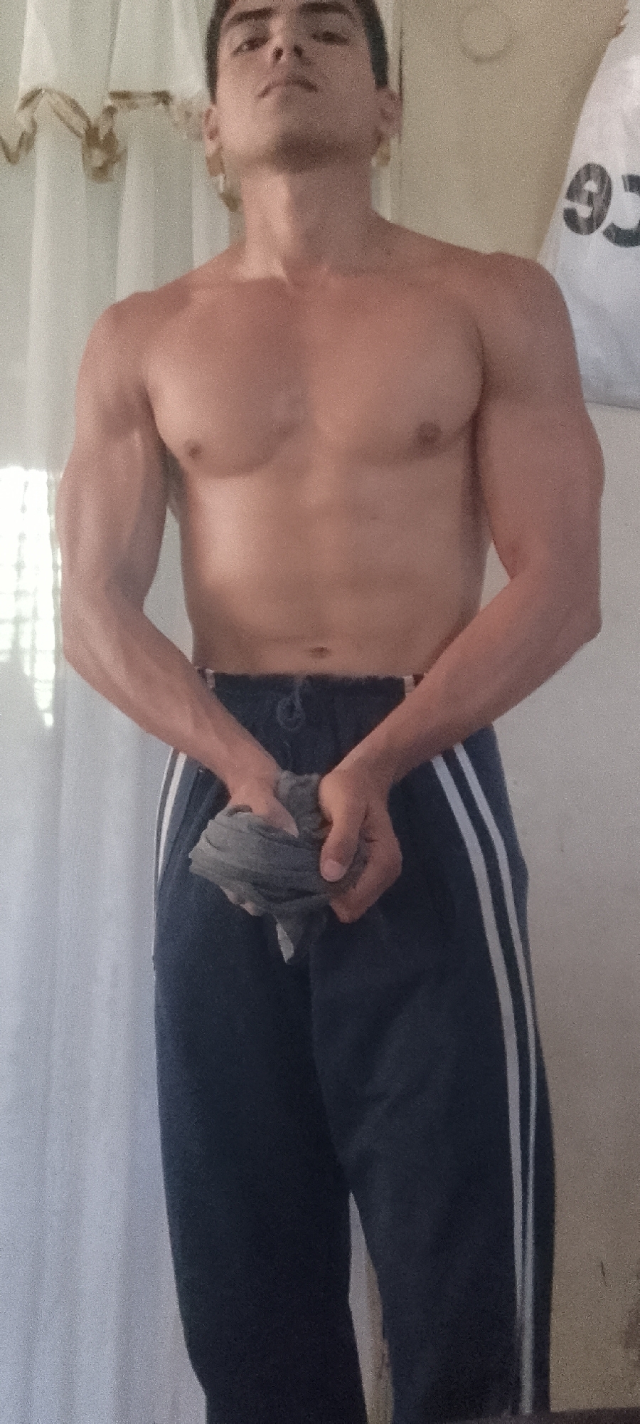 A shirtless man with dark hair and a muscular physique stands indoors, holding a grey cloth or towel in his hands. He is wearing dark blue track pants with white stripes down the sides. The background includes a light-colored curtain with a gold trim and a white bag or other item with the number "93" visible on it. No information about the location being Juan Griego, Venezuela is discernible from the image.