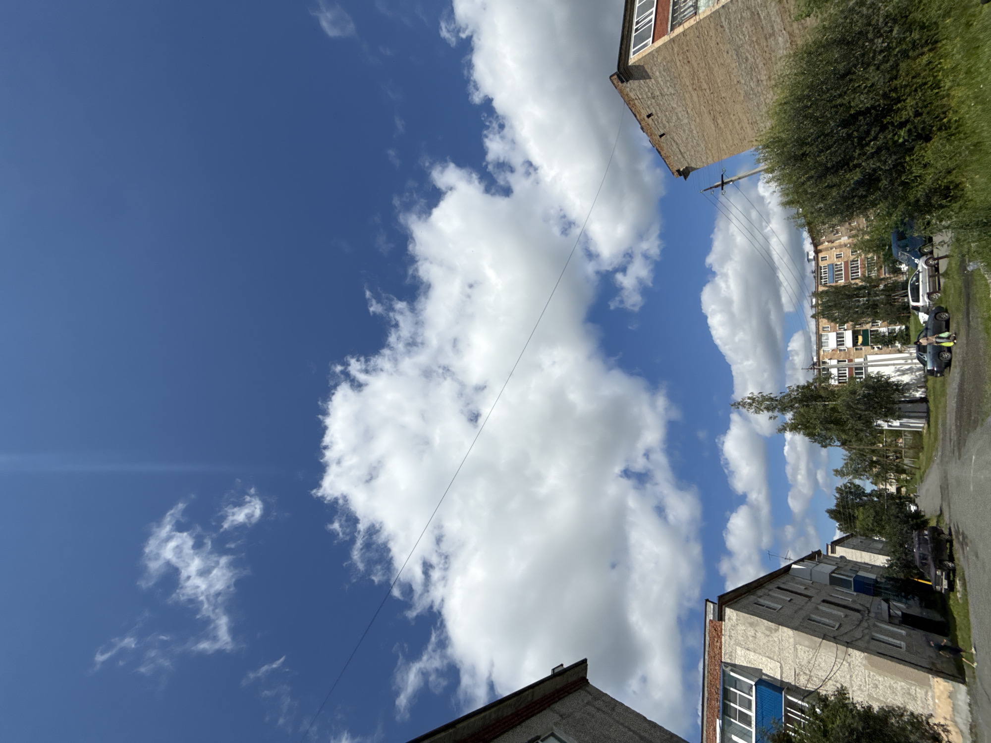A low-angle, wide-shot perspective of a street scene in Turinsk, Russia, under a bright blue sky with scattered white clouds. On the left, a gray, multi-story apartment building is angled dramatically, its facade showing multiple windows and balconies. In the foreground, a dark SUV is parked on a gravel road. Further down the street, other apartment buildings of similar architecture can be seen, along with several trees. To the right, a large brick building with visible window frames stands prominently. The overall impression is of a residential urban environment on a clear day.