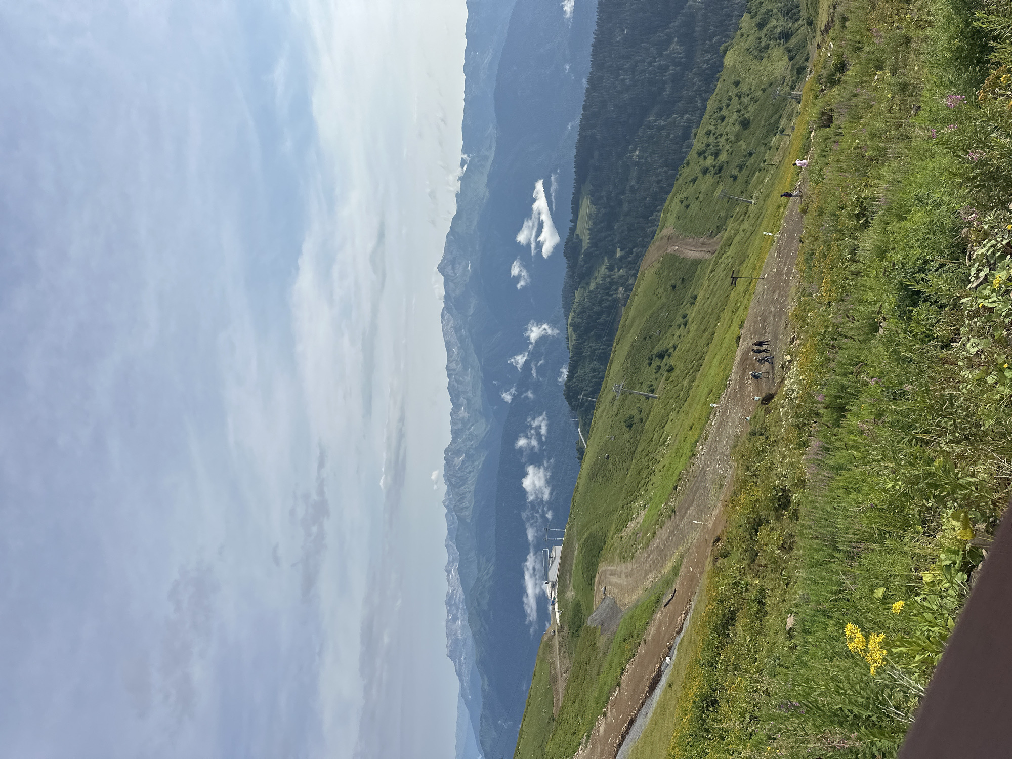 The image depicts a mountainous landscape with lush green slopes covered in vegetation and wildflowers. Several dirt paths wind across the slopes, and a few people are visible walking on them. In the mid-ground, a building and ski lift infrastructure are present. Further in the background, a range of majestic mountains stretches across the horizon, some capped with snow and shrouded in clouds. The sky is partly cloudy, with hues of blue and white. The scene appears to be from a ski resort area in Estosadok, Russia, likely taken during the summer or early autumn when the slopes are green but the higher peaks retain snow.