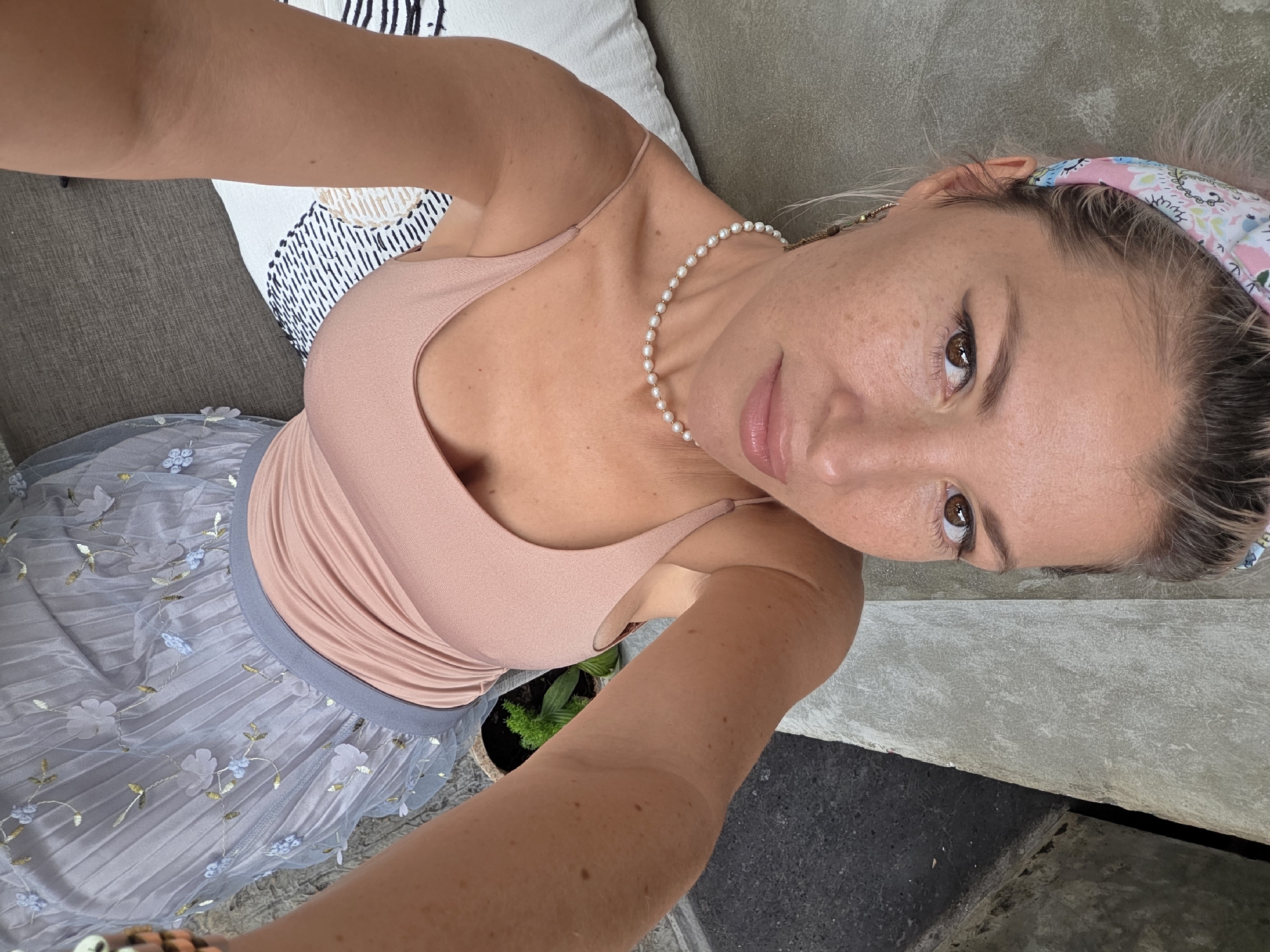 An overhead selfie taken by a woman with blonde hair tied back by a floral bandana, and light makeup. She is wearing a blush pink spaghetti strap top and a light blue pleated skirt with floral embroidery. A pearl necklace adorns her neck. She is seated on a gray sofa with a white and black patterned cushion. The background is a textured gray concrete wall and a dark concrete pathway. The location is likely a cafe or restaurant in North Kuta, Indonesia, given the style of the seating and the urban, outdoor setting.