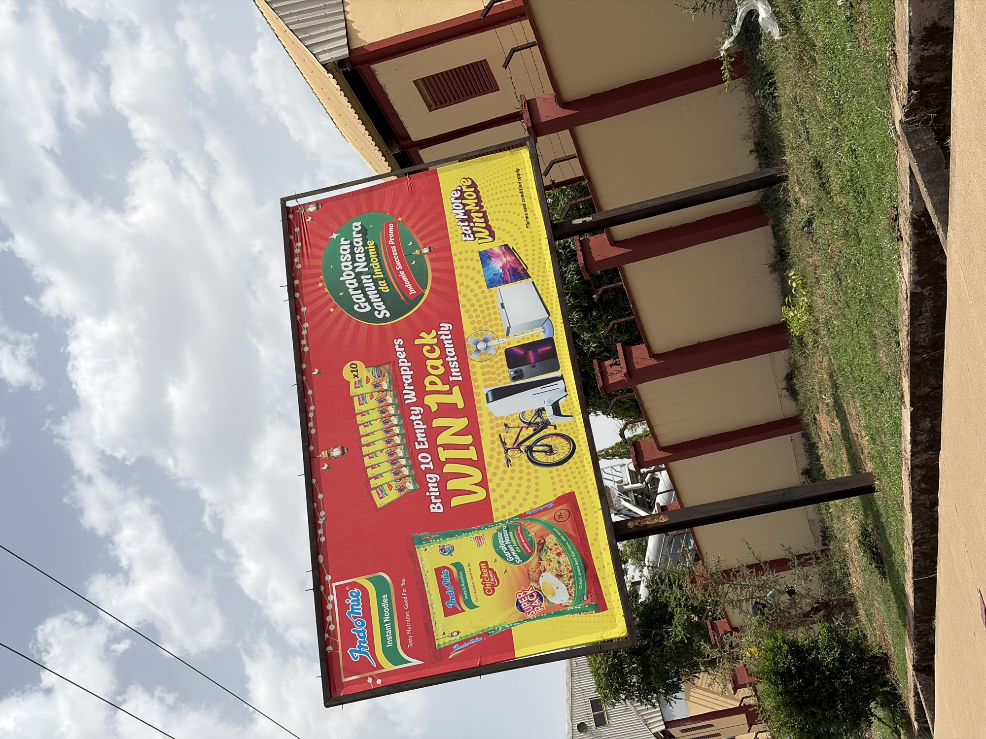 This media file is a rotated, outdoor shot primarily featuring a large, brightly colored billboard advertising Indomie instant noodles. The billboard, predominantly red and yellow, promotes a "Garabasar Samun Nasara da Indomie" (likely Hausa for "Indomie Success Promo") with the slogan "Eat More, Win More." A key part of the promotion is visible: "Bring 10 Empty Wrappers WIN 1 Pack Instantly." The billboard displays various potential prizes, including a flat-screen television, a refrigerator, a smartphone, and a bicycle. Images of Indomie noodle packets, specifically "Chicken Flavor" and a "SUPER PACK," are prominent, alongside text such as "Indomie Instant Noodles" and "Tasty Nutrition. Good For You." A cluster of ten small noodle packets is also shown, labeled "x10." A disclaimer "*Terms and condition apply" is also present.
The setting appears to be an open area in Rafin Par, Nigeria, under a cloudy, daytime sky with power lines visible. In the background, behind the billboard, are structures with yellow walls and red support beams, alongside green grass and some sparse vegetation. No people or specific activities are visible, focusing solely on the advertisement and its immediate surroundings.