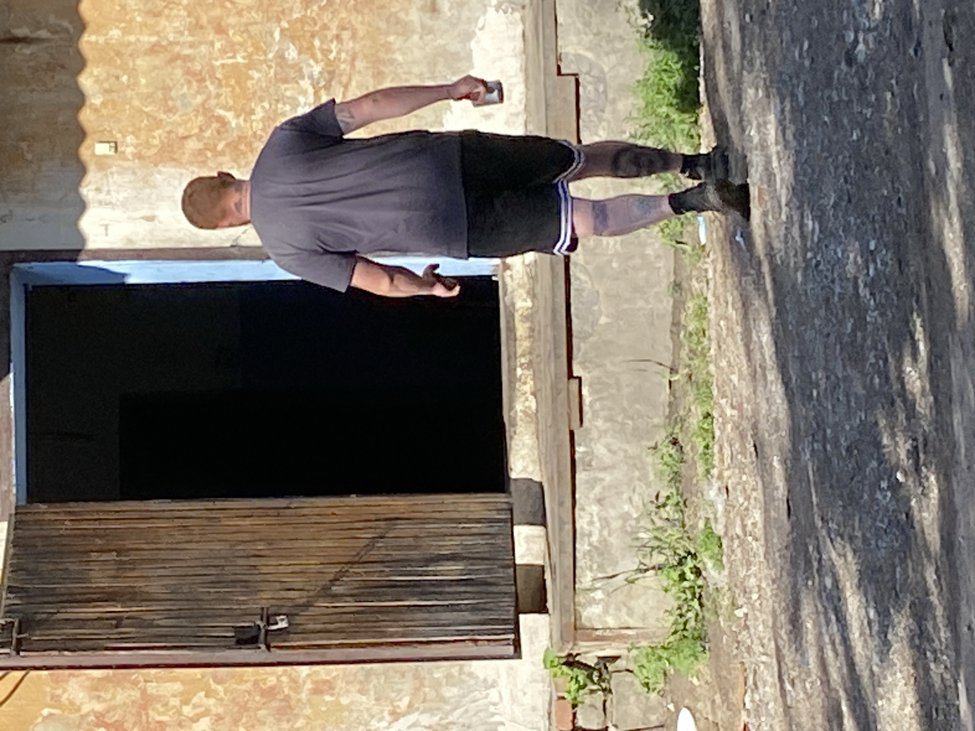 A man with red hair and tattoos on his arms and legs walks away from the camera towards an open doorway. He is wearing a dark t-shirt, black shorts with white and blue stripes, and dark boots. He holds a small cup in his right hand. The man is walking on a gravel path in front of a weathered, textured wall. To his left, a large wooden door is ajar. The environment appears to be outdoors, possibly in a rural or industrial setting, with some overgrown vegetation visible near the wall. The lighting suggests it is daytime, with shadows cast on the ground.