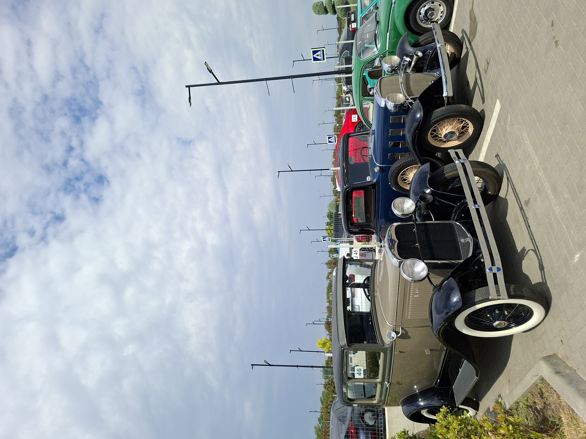 The image shows a row of vintage cars parked in an outdoor lot. The cars are from different eras, with the most prominent being a light brown and black Ford Model A in the foreground. Behind it are a dark blue car, a green car, and a portion of another vehicle, all appearing to be older models. The cars are parked on a paved surface with white lines indicating parking spaces. In the background, there are streetlights, some trees, and other vehicles parked in the distance. The sky is partly cloudy with a mix of blue sky and white clouds. A sign with the number "46" is visible on the side of the brown car. The scene appears to be in a public parking area. The location is Stăuceni, Moldova.