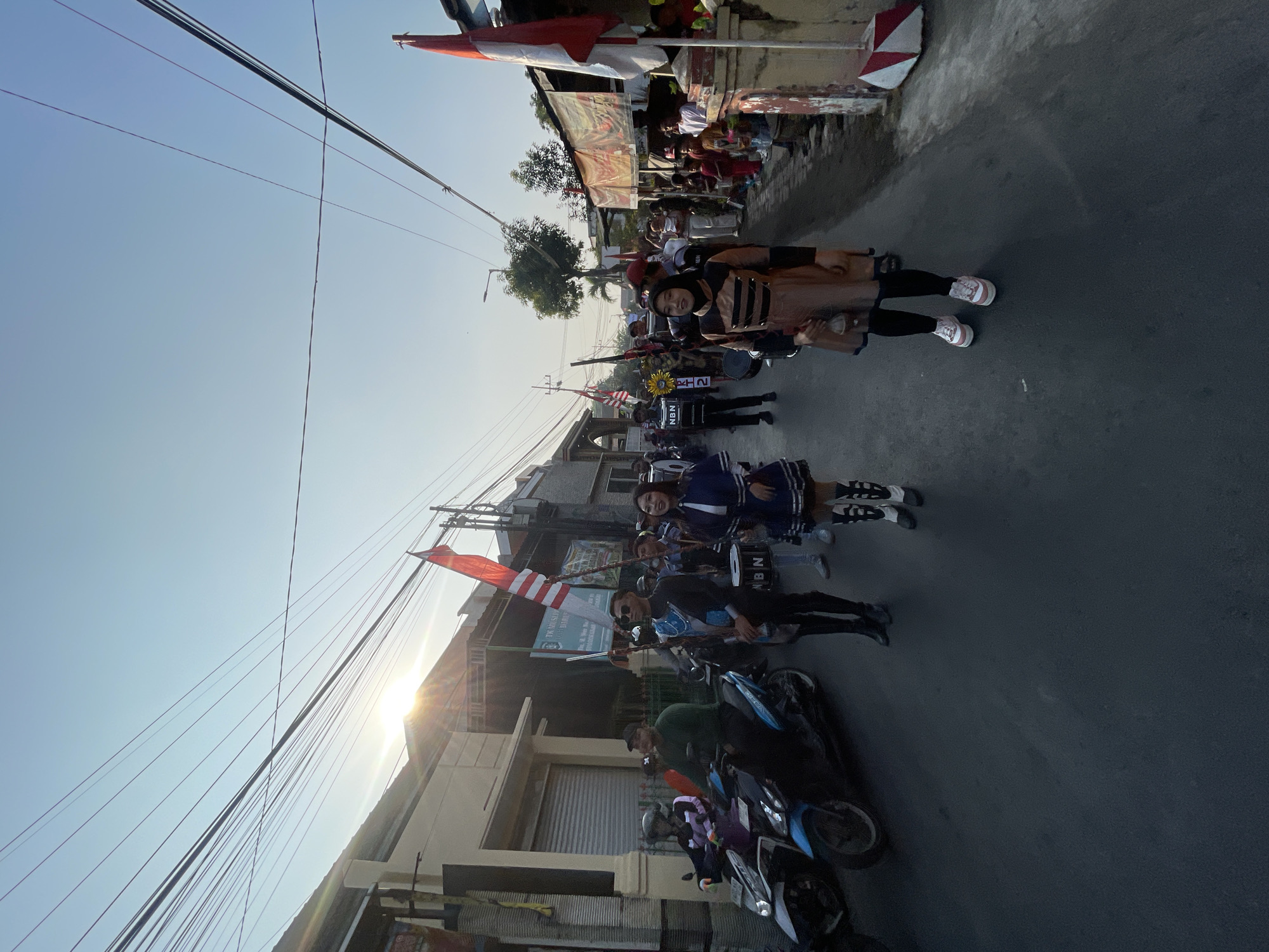 The image depicts a street scene in Candi, Indonesia, likely during a parade or celebratory event. The foreground features several individuals in uniform, possibly part of a marching band. One person on the left is wearing a black uniform with blue accents and carrying a flag. Next to them, a young woman in a blue and white pleated dress with tall boots is walking. Further to the right, another individual in a brown and black outfit with a skirt and black leggings, also wearing sneakers, is visible. In the background, there are buildings lining the street, some with banners and flags. Several motorcycles are parked or moving on the side of the road. The sun is setting or rising, casting a warm glow and creating lens flare. The sky is clear and blue, with a complex network of overhead electrical wires crisscrossing the frame. The street itself is paved with asphalt. The overall atmosphere appears festive and community-oriented.