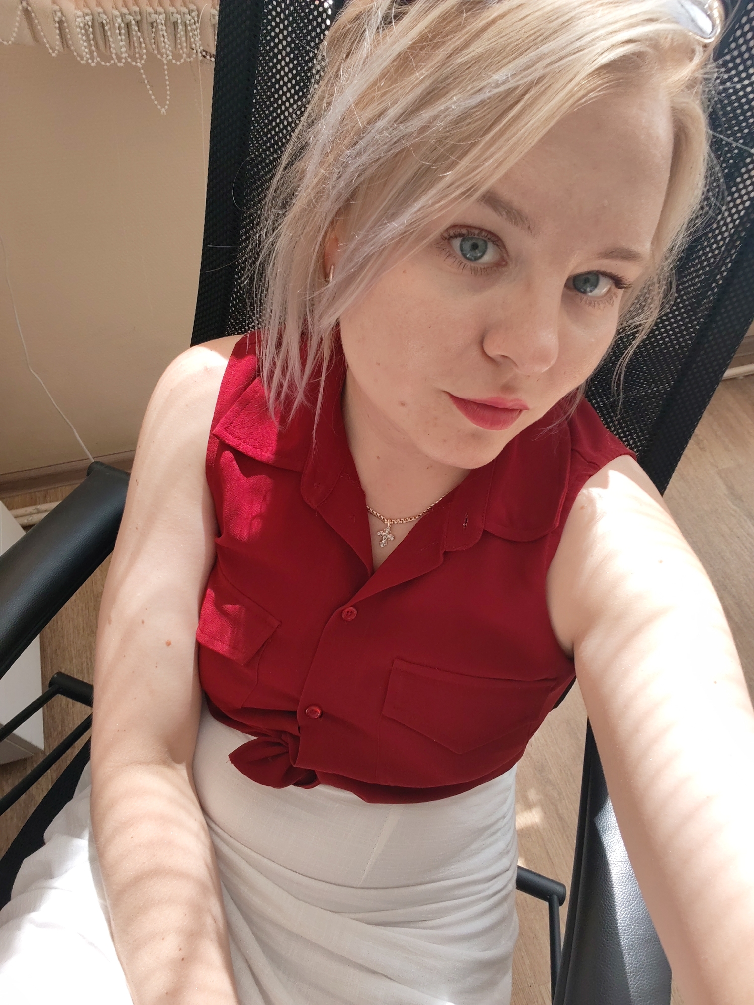 A fair-skinned woman with blonde hair and blue eyes is seated in a black office chair indoors. She is wearing a red, sleeveless, collared shirt with two small pockets on the chest, buttoned partially down the front, and tied at the waist, paired with a white skirt. She wears a gold necklace with a cross pendant and small hoop earrings. The room has light-colored walls and a wooden floor. A beaded curtain hangs in the background. The exact location in Izhevsk, Russia, is indeterminable from the given information.