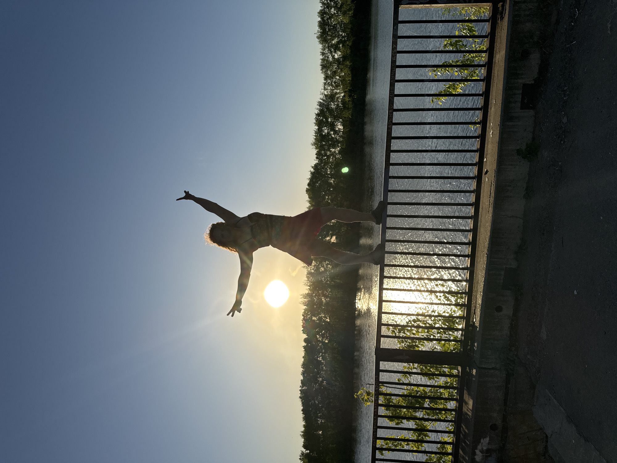 The image shows a person striking a yoga pose on a concrete structure next to a body of water, with a metal railing separating them from the water. The sun is setting behind the person and the water, creating a silhouette effect. The person is wearing a patterned top and red shorts. Trees can be seen in the background on the opposite side of the water. Based on visual clues, the location is Chaikovsky, Russia. The person appears to be interacting with the environment by posing in front of the sunset.