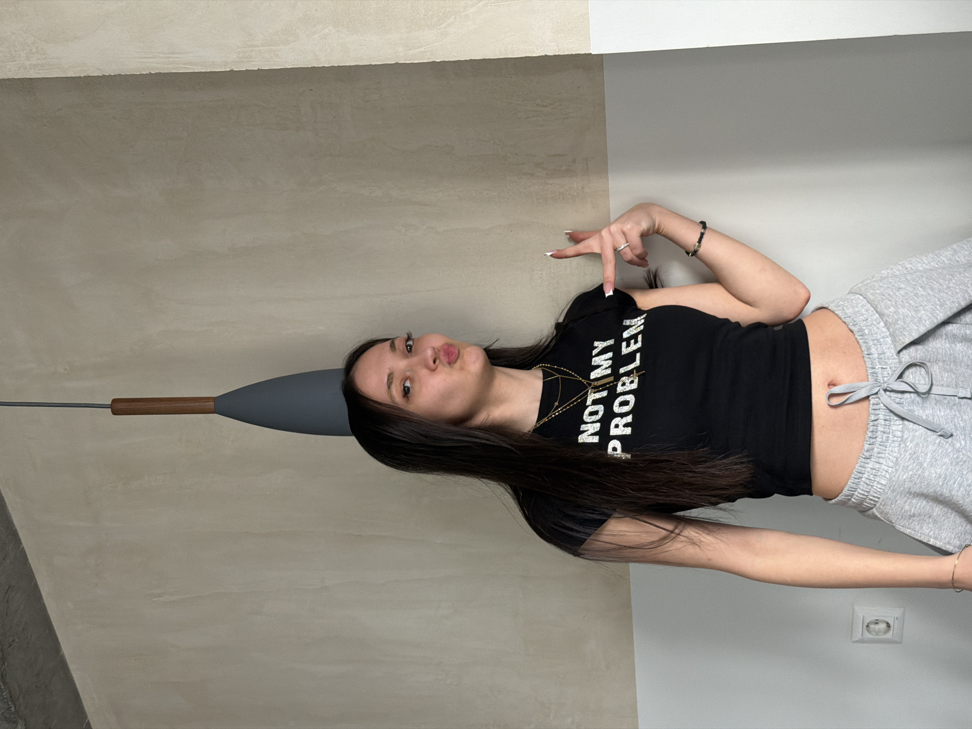 A woman stands indoors. She wears a black t-shirt with "NOT MY PROBLEM" in white lettering, gray sweatpants, and jewelry. She has dark hair and fair skin. Her left arm is raised with fingers extended in a "peace" sign, while her right arm is at her side. The background includes a wall with a gray area on the left and a white area on the right. A light fixture with a gray cone and wooden handle extends from the gray area. An electrical outlet is visible on the white wall near her right side. The location of the photograph is not stated to be in the city of Yerevan, Armenia.