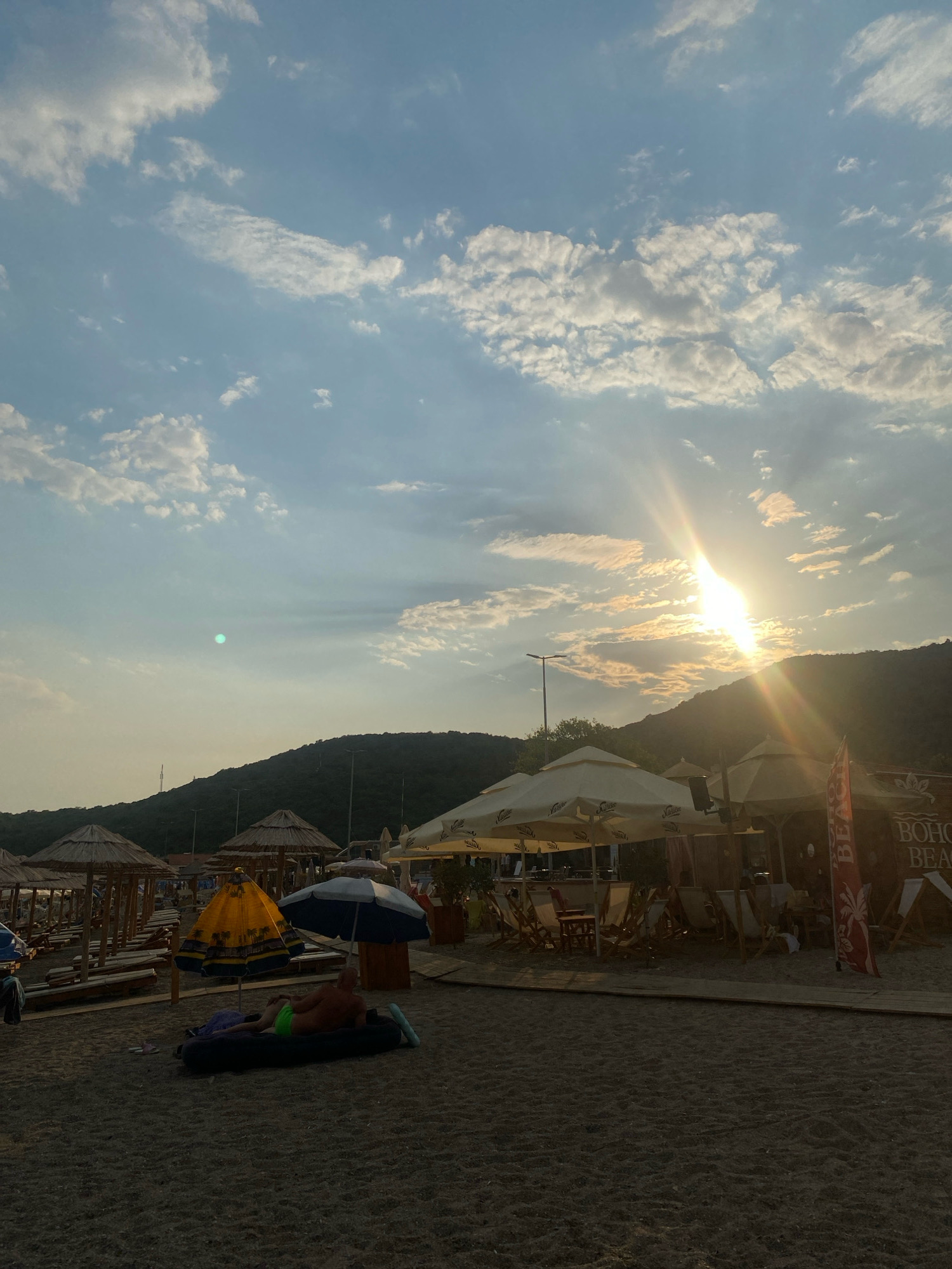 The image depicts a beach scene in Prijevor, Montenegro, during what appears to be late afternoon or early evening. The sun is setting behind a lush, green mountain range that forms the backdrop. Its rays are prominently featured, breaking through the partly cloudy sky and casting a warm glow over the scene.
In the foreground, there is a sandy beach with rows of lounge chairs and thatched umbrellas, suggesting a resort or public beach area. A man is lying on an inflatable mattress on the sand, wearing green swim trunks. He is facing away from the camera, seemingly relaxing.
Several beach umbrellas are visible, including a distinctive yellow one with a patterned design and a blue and white one. To the right of the image, there's a structure that appears to be a beach bar or cafe, with tables and chairs set up under large white umbrellas. A red flag with white lettering, possibly indicating the name of the establishment, is positioned near the bar.
The sand is a dark, granular texture, and the overall lighting suggests a peaceful and idyllic atmosphere as the day draws to a close. The sky is a mix of blue with scattered white and grey clouds, some of which are illuminated by the setting sun.