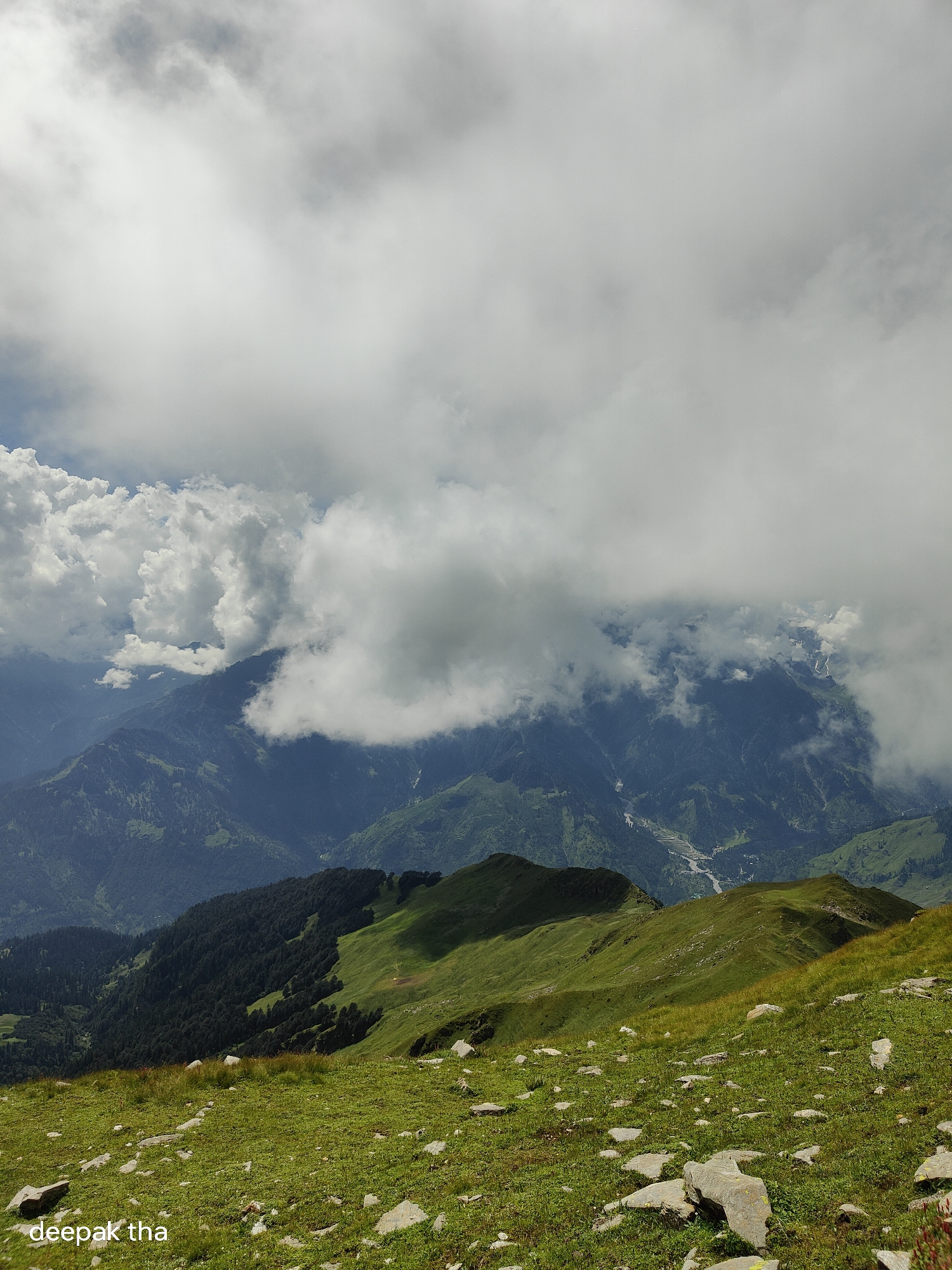 The image depicts a mountainous landscape with lush green slopes and rocky terrain in the foreground. A dense forest covers the lower slopes of the mountains in the mid-ground. The background features more rugged mountain peaks partially obscured by dramatic, swirling clouds, suggesting a high-altitude location. A river or valley can be seen winding through the distant mountains. The sky is filled with a mixture of bright white and darker grey clouds, indicating dynamic weather conditions. A watermark "deepak tha" is present in the bottom left corner. The scene is an outdoor, natural environment.