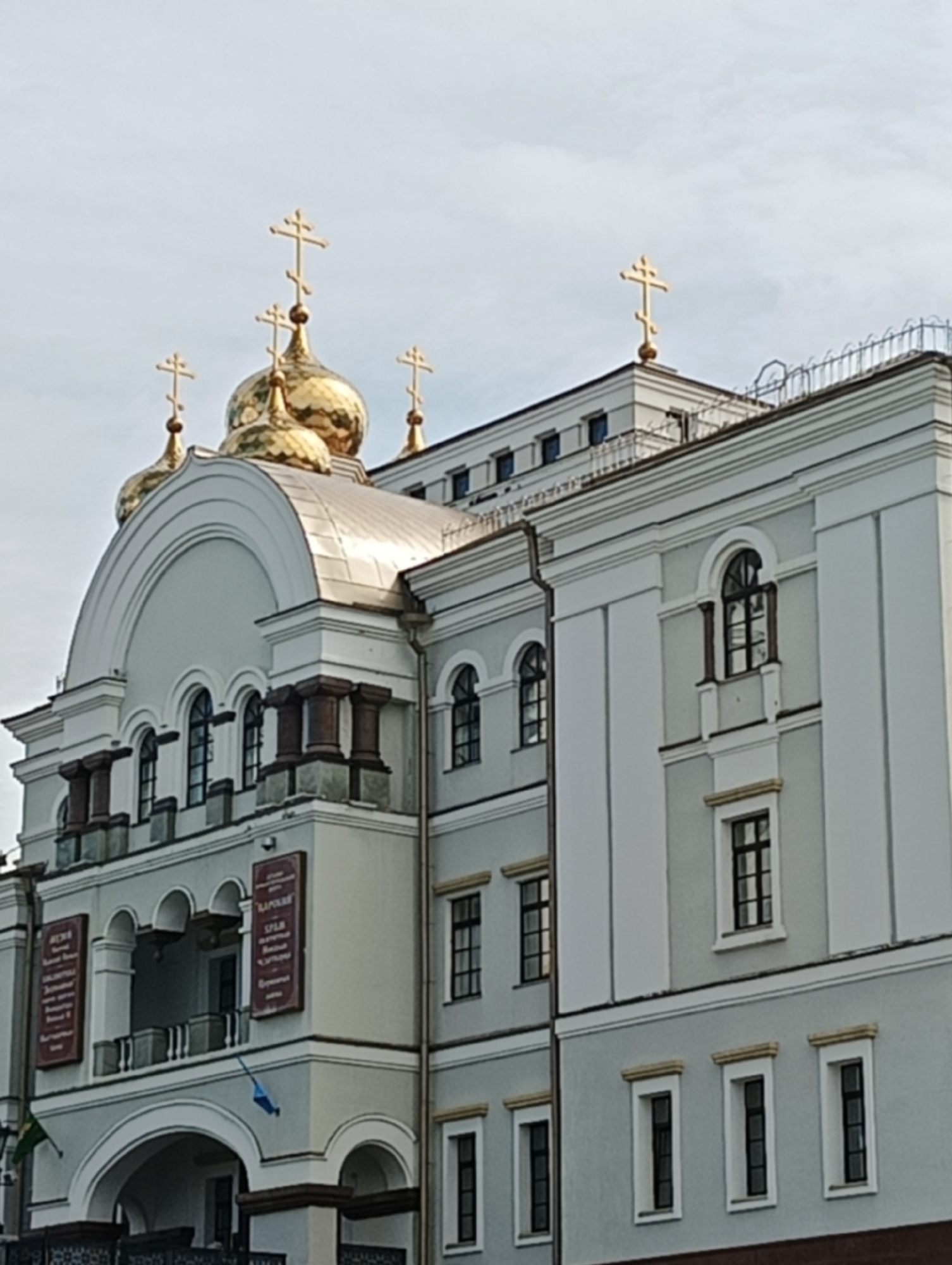 The image depicts a building in Yekaterinburg, Russia, identifiable as an Orthodox church based on its architectural style. The structure has multiple golden domes, each topped with a cross. The building's facade is primarily light gray, with white and dark brown accents. The architecture incorporates arched windows and decorative columns. Signboards with inscriptions are visible on the building's exterior. A small flag hangs near the entrance. The sky is overcast, providing diffused lighting. There are no people visible in the image. The building appears well-maintained and is likely a landmark in the city.
