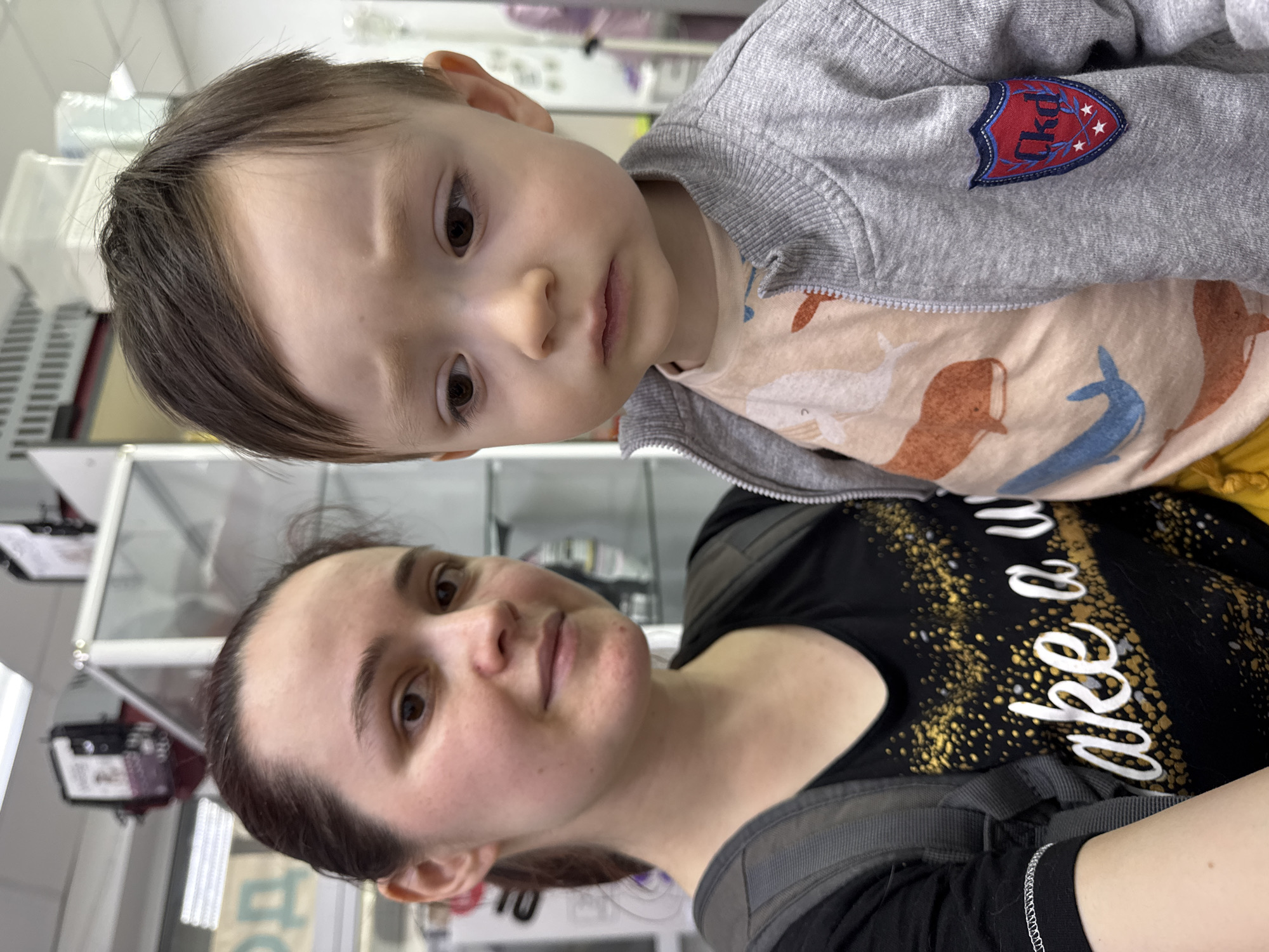 A woman holds a young child in her arms. The woman has brown hair pulled back, light skin, and is wearing a black t-shirt with white and gold lettering. She has a gray backpack on. The child has brown hair, light skin, and is wearing a gray zip-up jacket over a printed t-shirt. The background includes what appears to be retail display cases, potentially inside a store. The location may be Izhevsk, Russia.